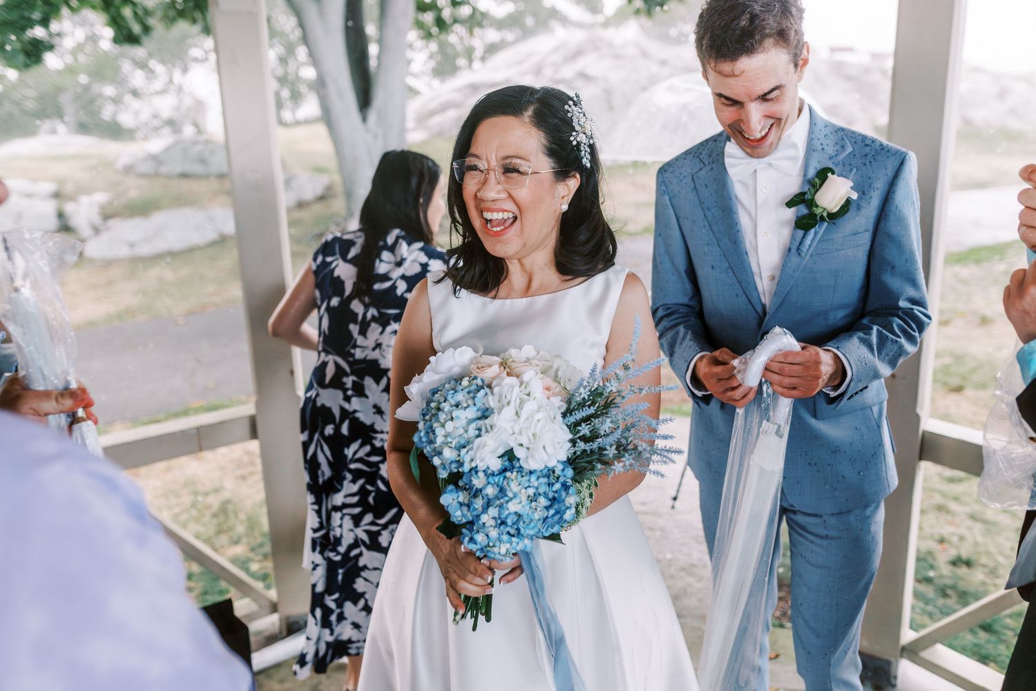 Wedding party shares joyful moment under white gazebo overlooking coastal scenery.