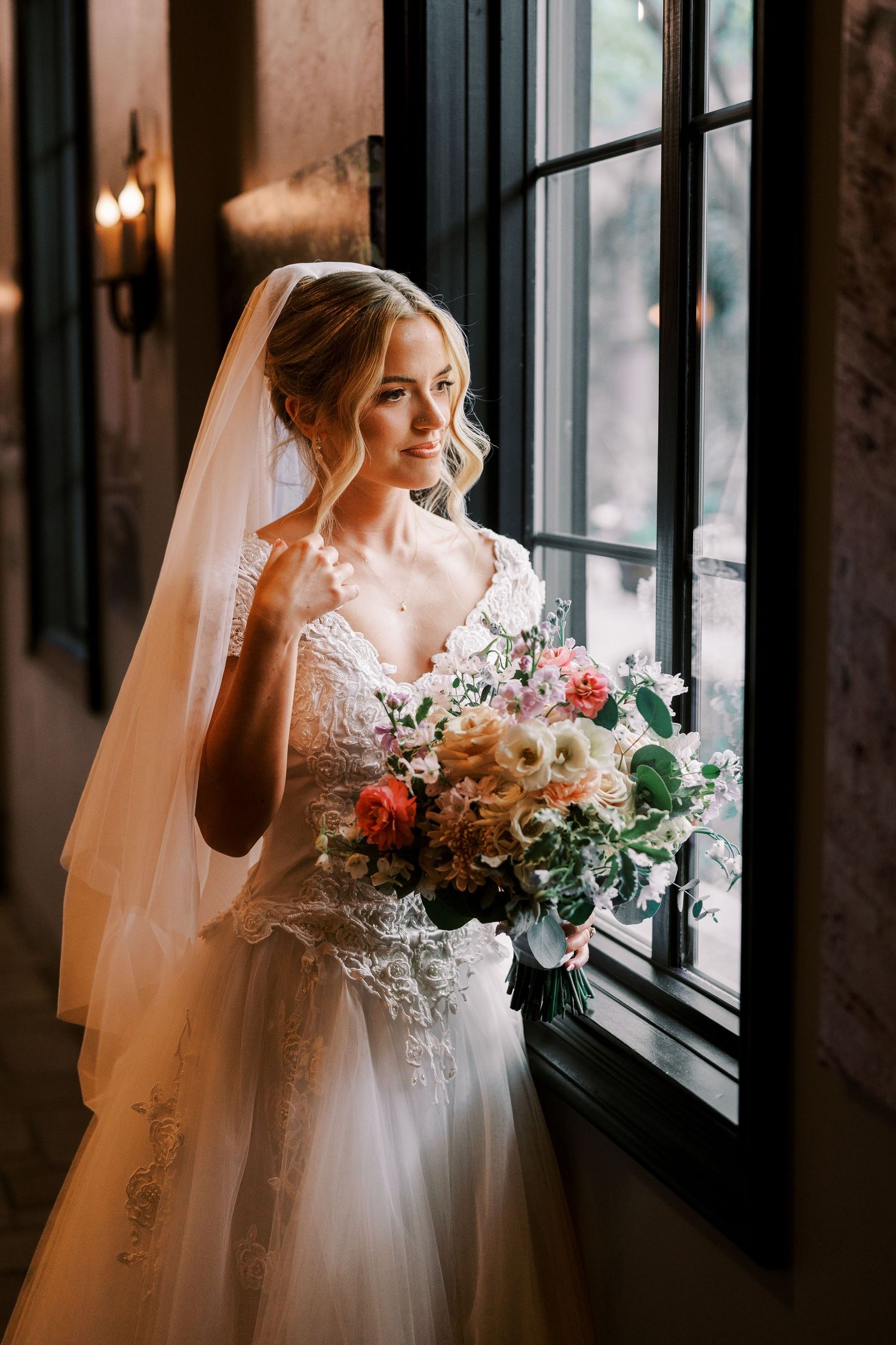 A bride in a white lace gown holds a romantic peach and coral bouquet while standing by a window in moody lighting.