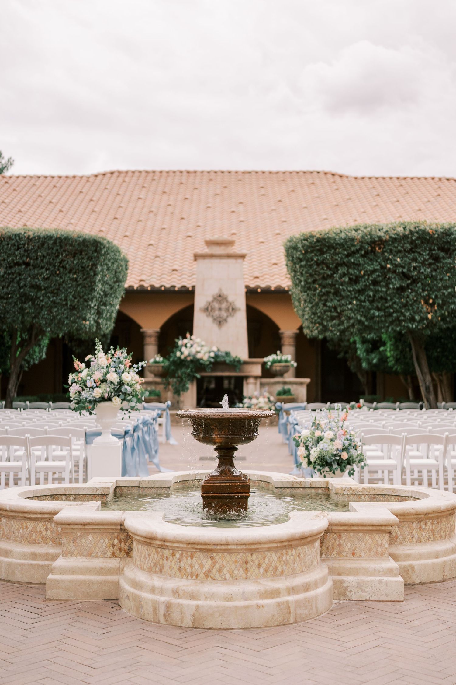 Elegant outdoor wedding venue with stone fountain and white chairs arranged around curved stone walls at sunset.