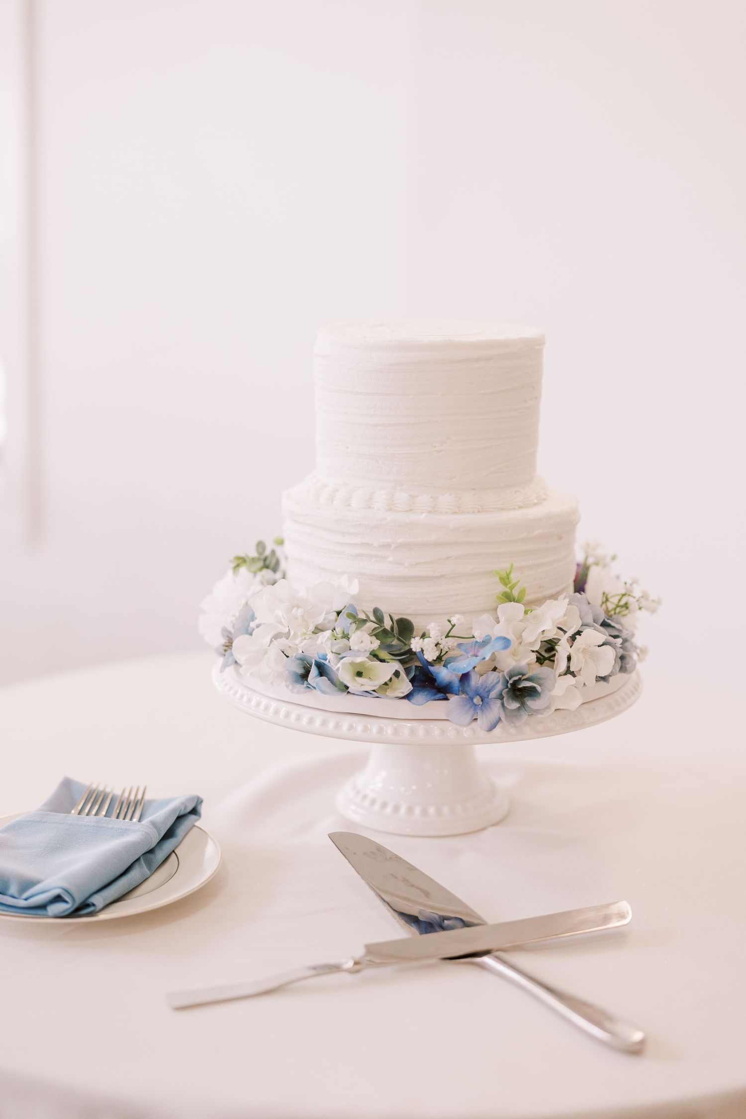 A minimalist white wedding cake with delicate blue floral accents displayed on a white cake stand with table settings.