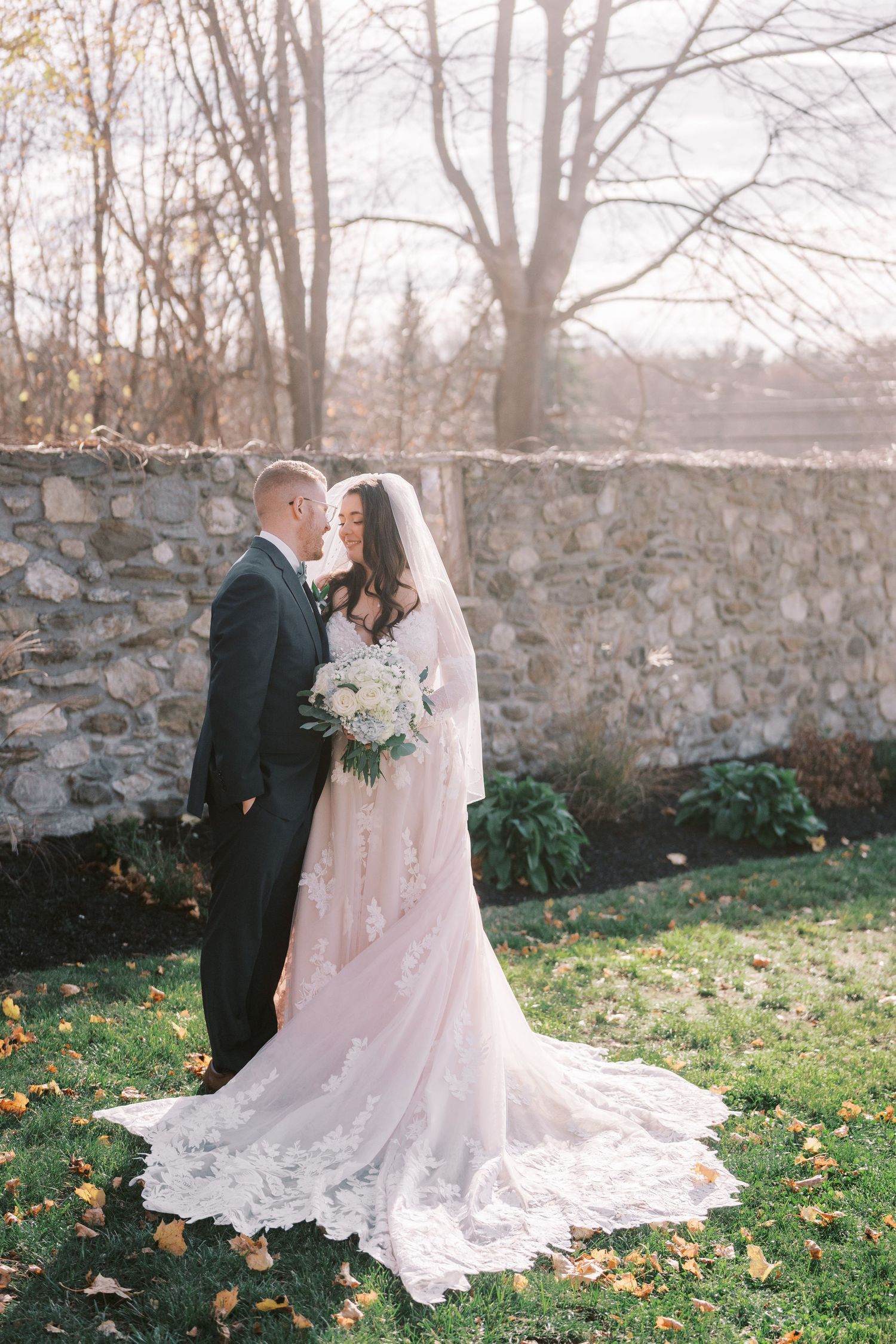 A bride and groom share an intimate moment against a rustic stone wall with autumn leaves scattered on the ground.