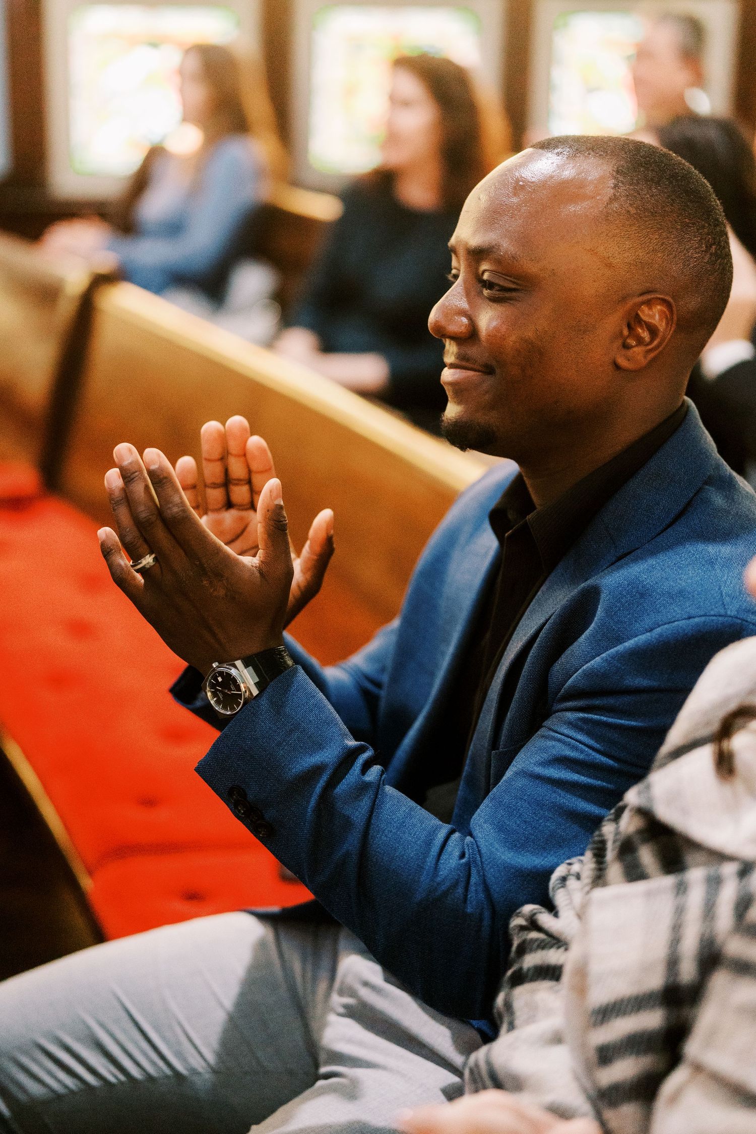Wedding guest in a blue blazer laughs expressively while seated in church pews.