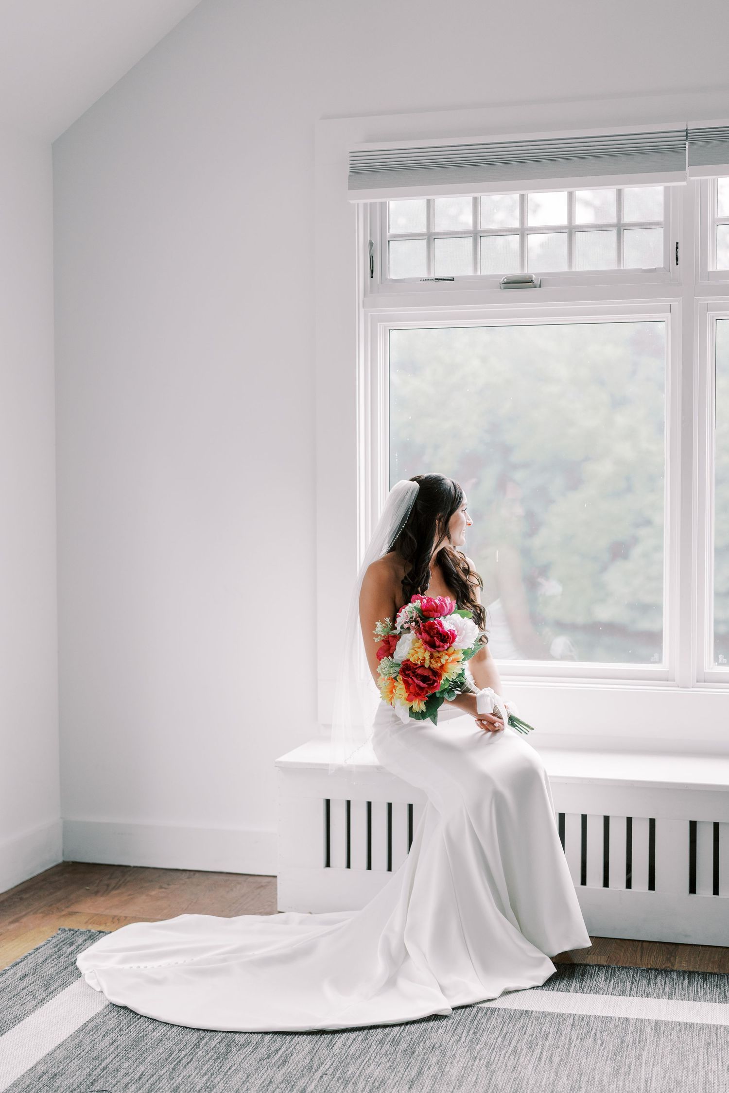 A bride in a flowing white dress poses gracefully near a window in a bright minimalist room with hardwood floors.