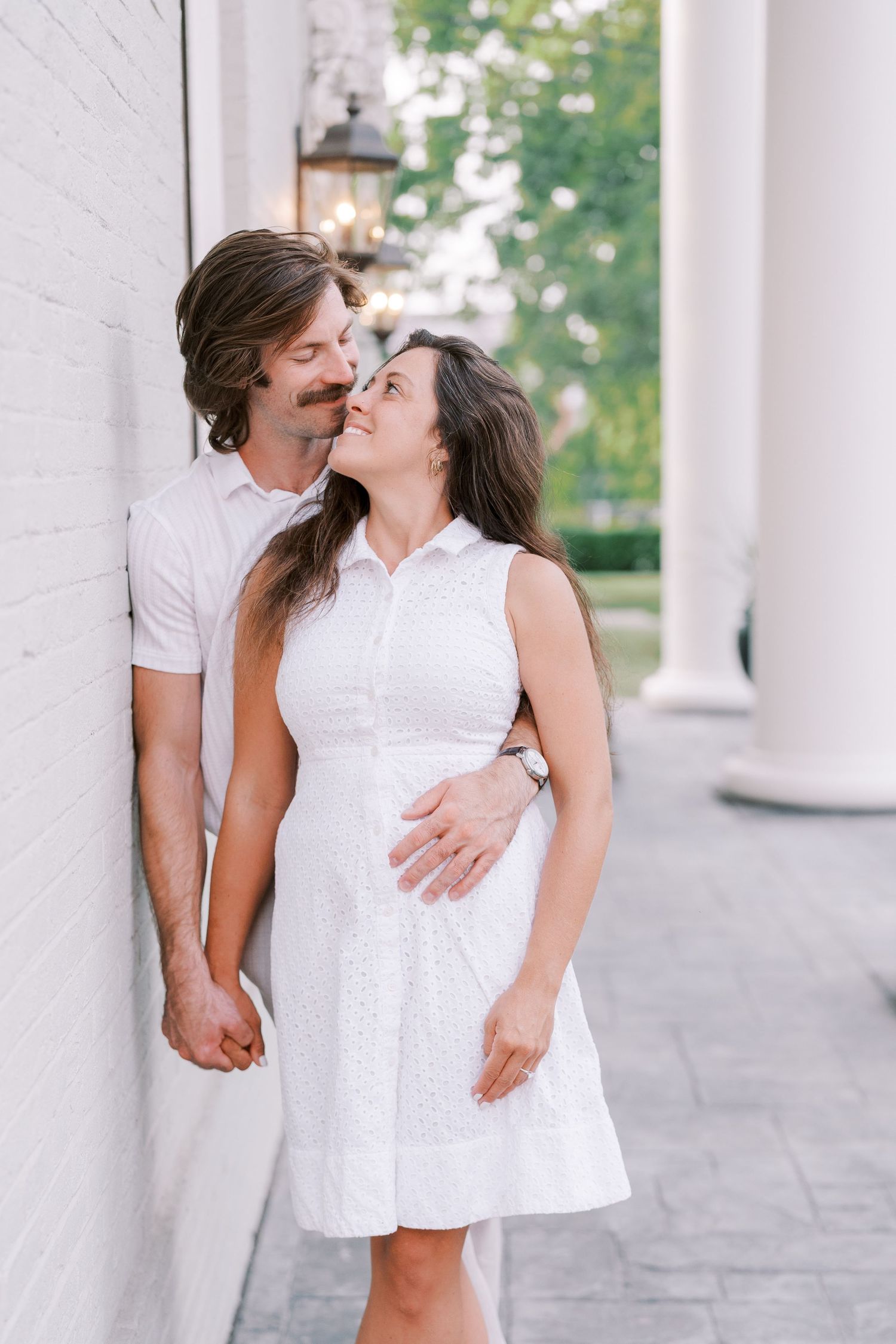 A couple in white outfits share intimate moments against white columns outdoors.