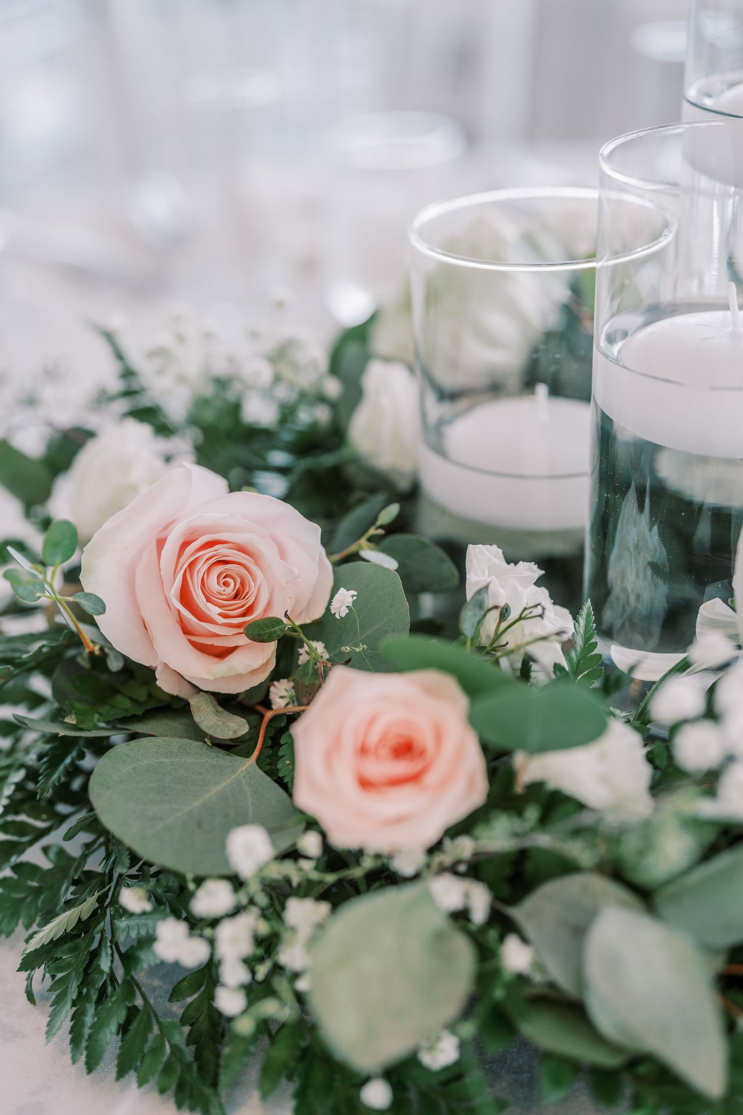 Delicate pink roses and baby's breath arranged with eucalyptus leaves around glass candle holders in an elegant centerpiece.