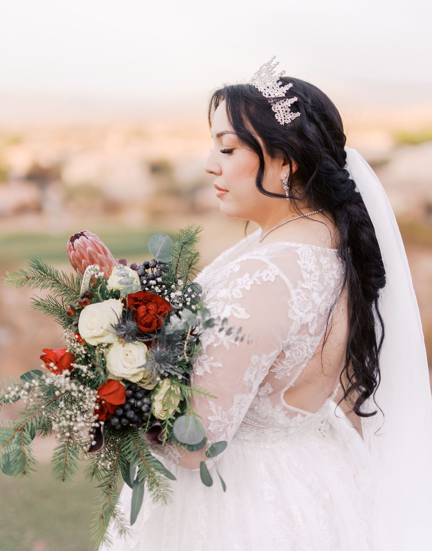 A bride in a lace dress with open back detail holds a romantic burgundy and white bouquet at sunset.