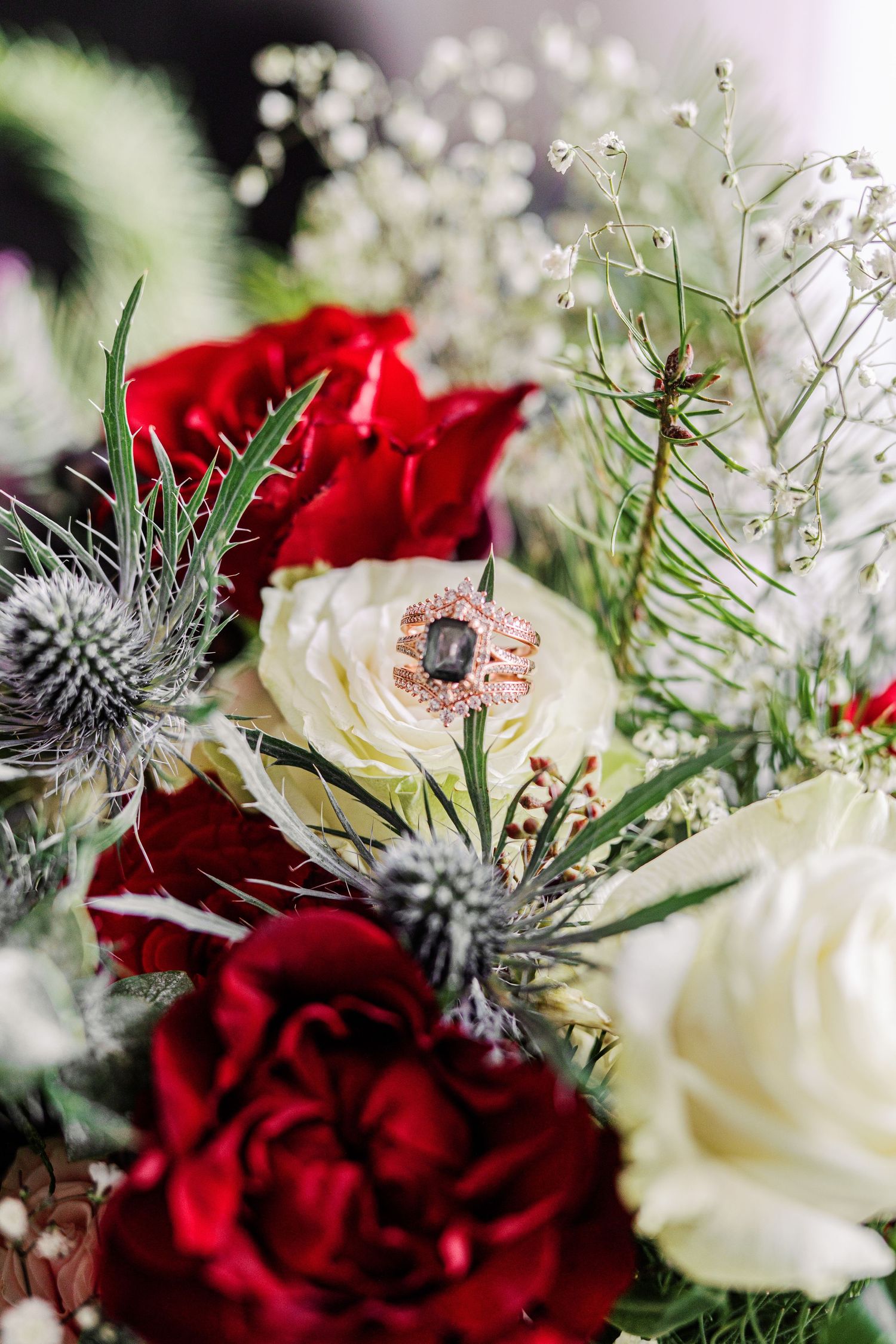 A rose engagement ring rests among red and white roses with blue thistles and baby's breath in a bridal bouquet.