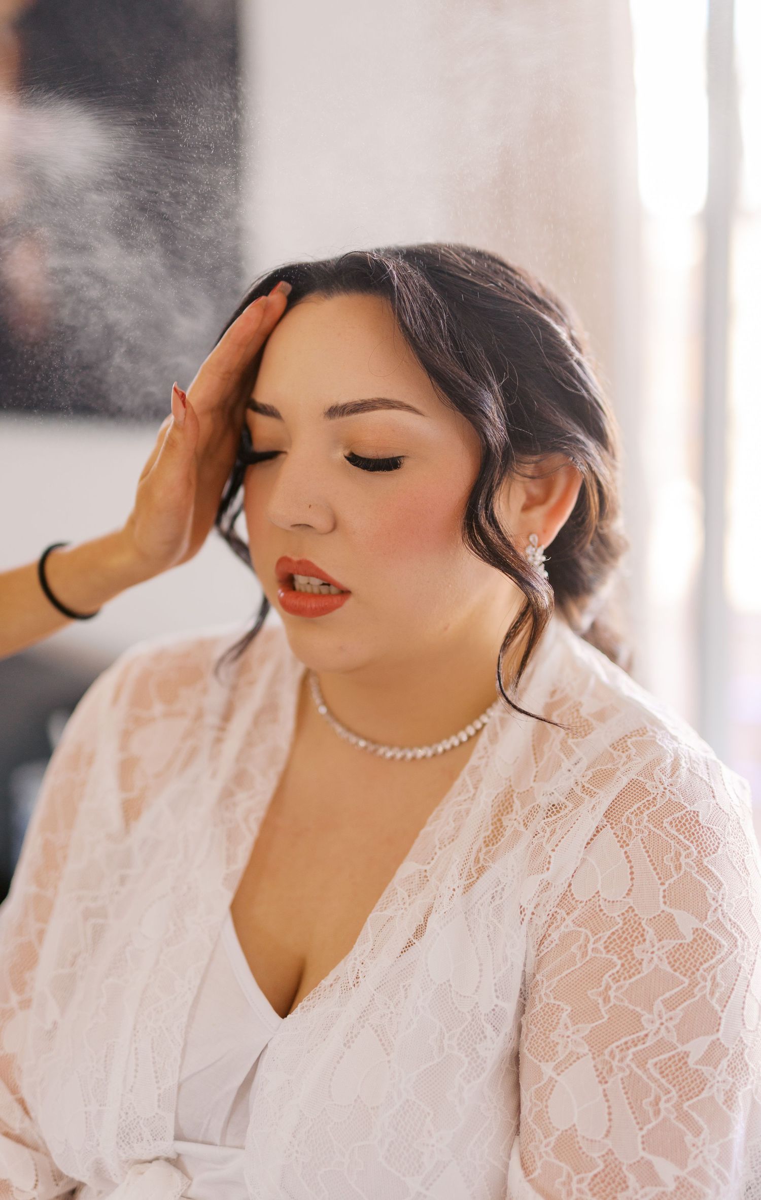 A person wearing a delicate white lace dress and pearl necklace gets ready with eyes closed in soft natural light.