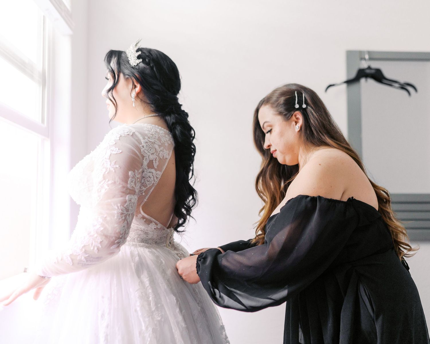 Wedding preparations show a tender moment as an attendant helps adjust a bride's flowing white lace gown by a window.