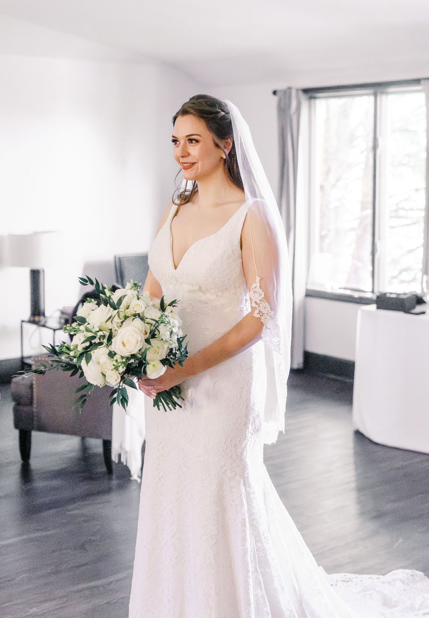 Bride in white lace gown poses in a bright modern room with grey floors and white walls.