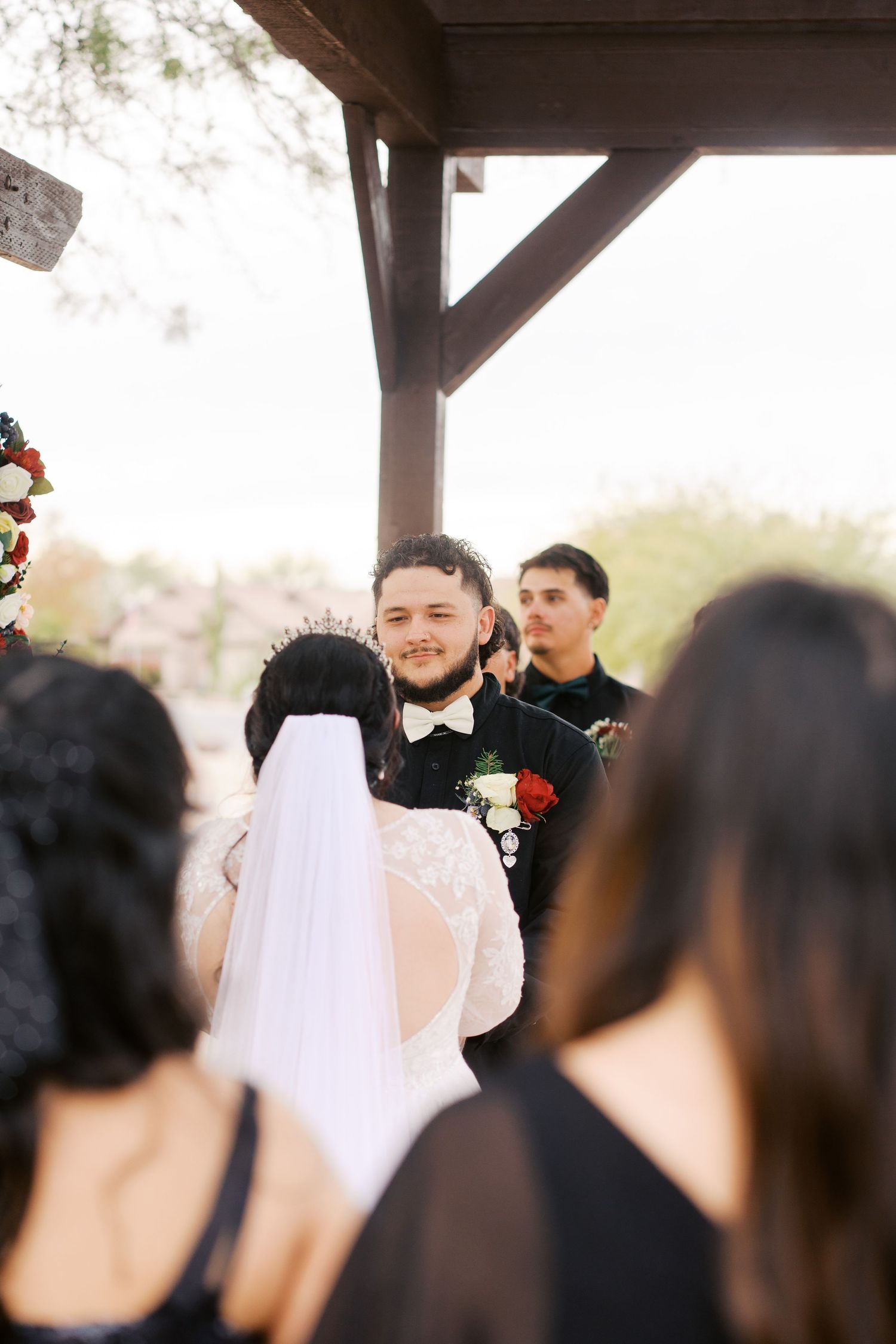A wedding ceremony takes place under a wooden pergola with the couple and guests gathered during an outdoor celebration.