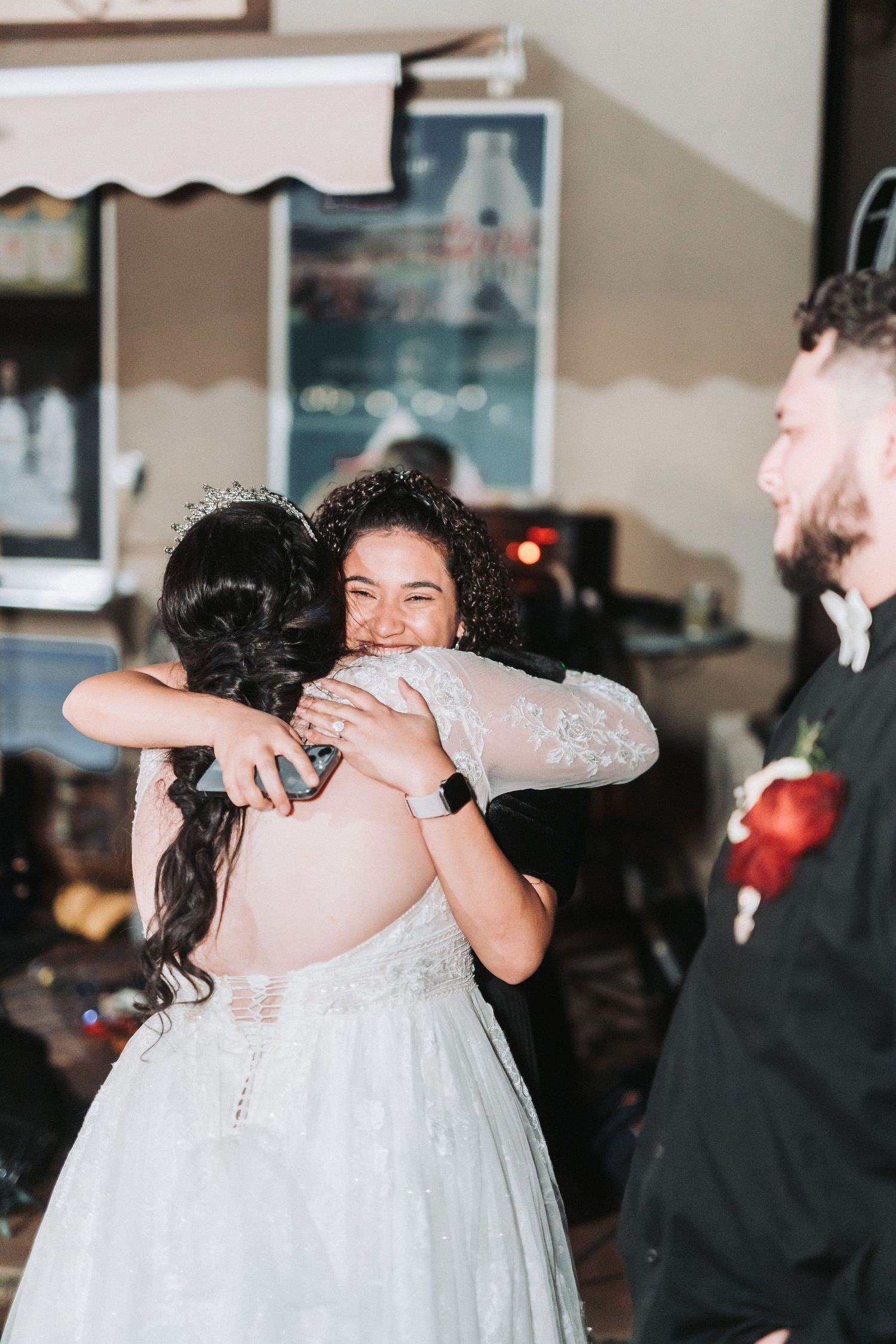 Two people share an emotional embrace at a wedding reception while a guest in a black suit looks on.