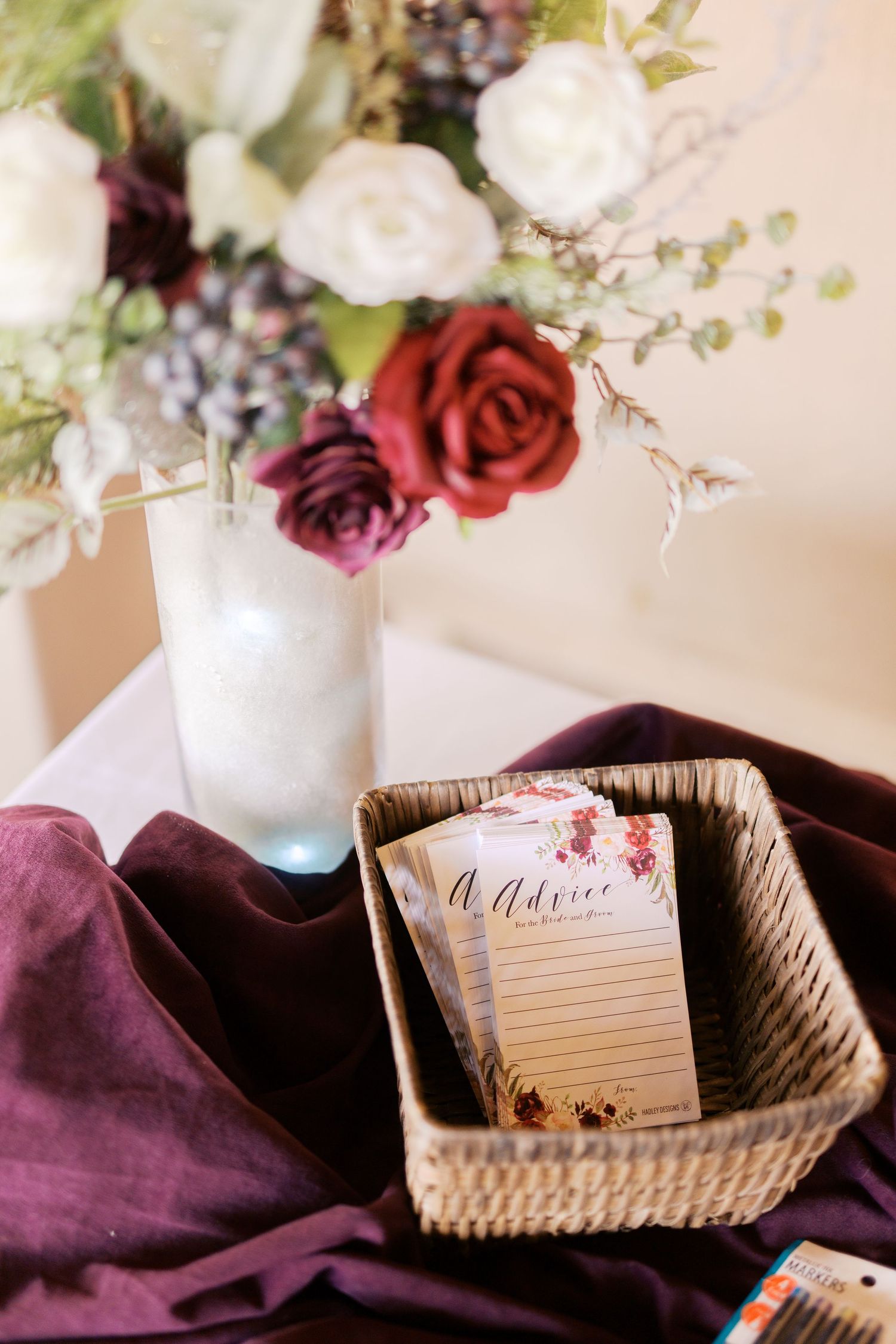 A floral notepad sits in a woven basket beside white and burgundy roses in a frosted vase with purple fabric.