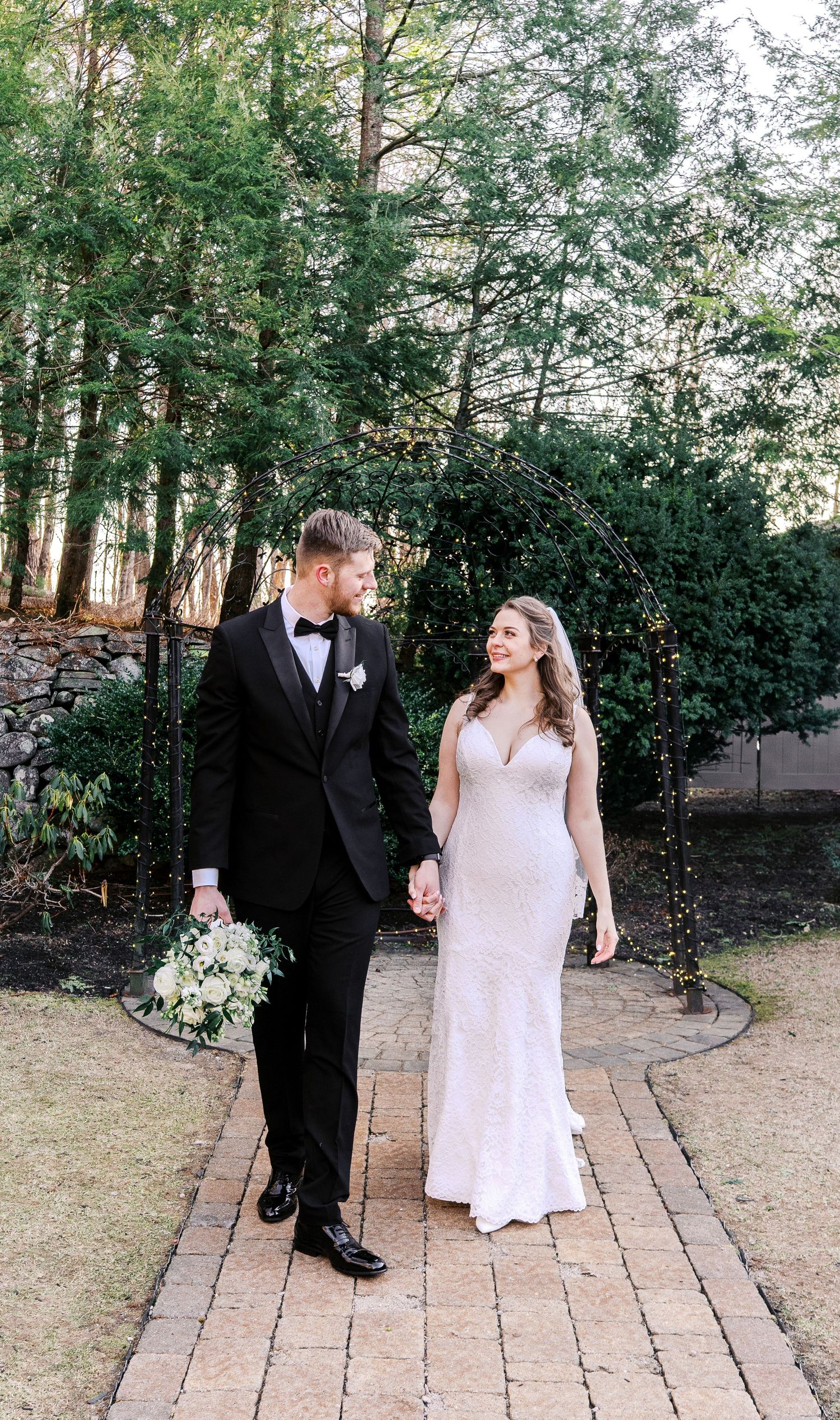 Romantic couple strolling along a brick pathway surrounded by lush greenery and trees.