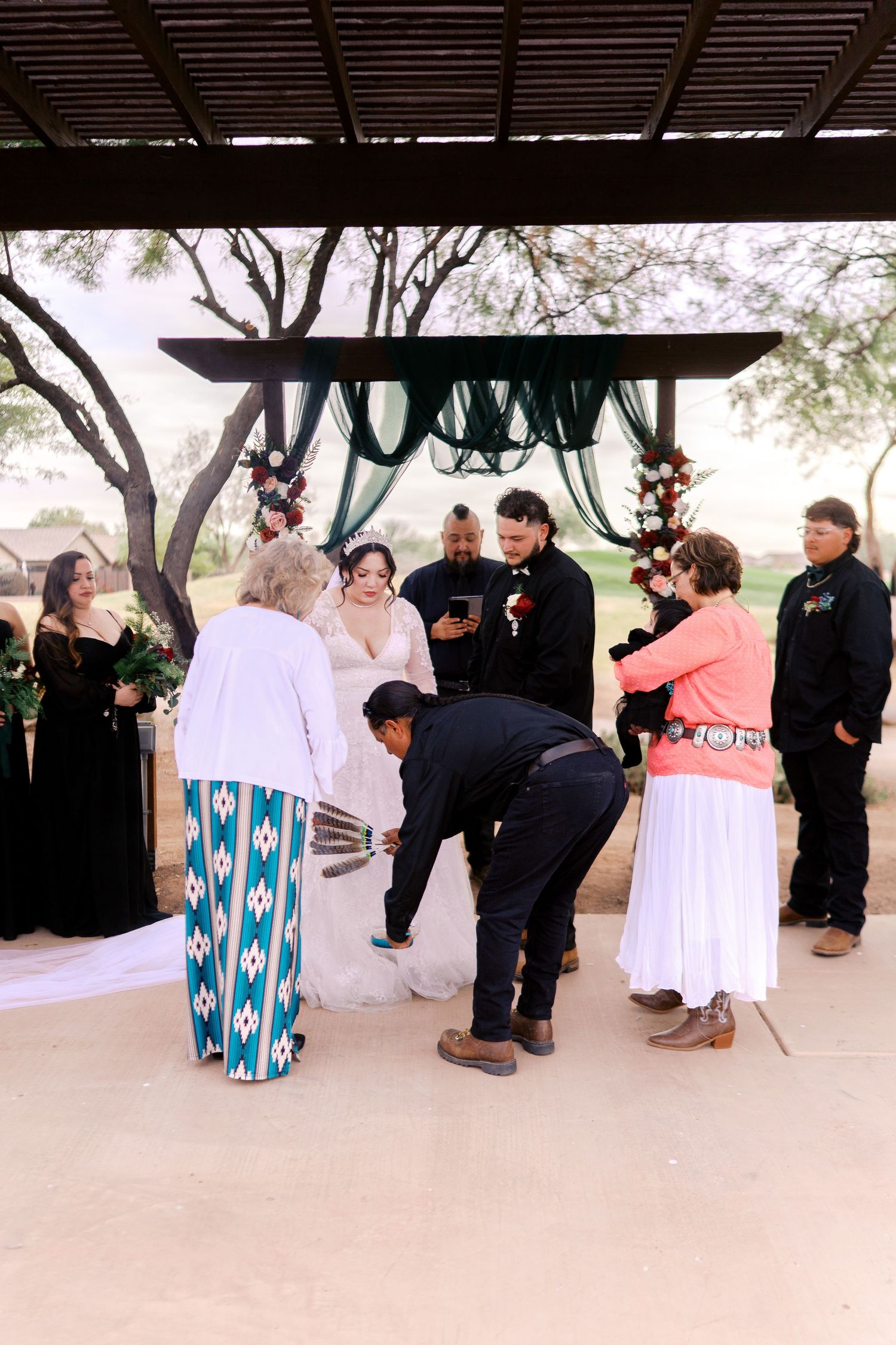 A wedding ceremony takes place under a wooden pergola with turquoise decor and gathered guests outdoors.
