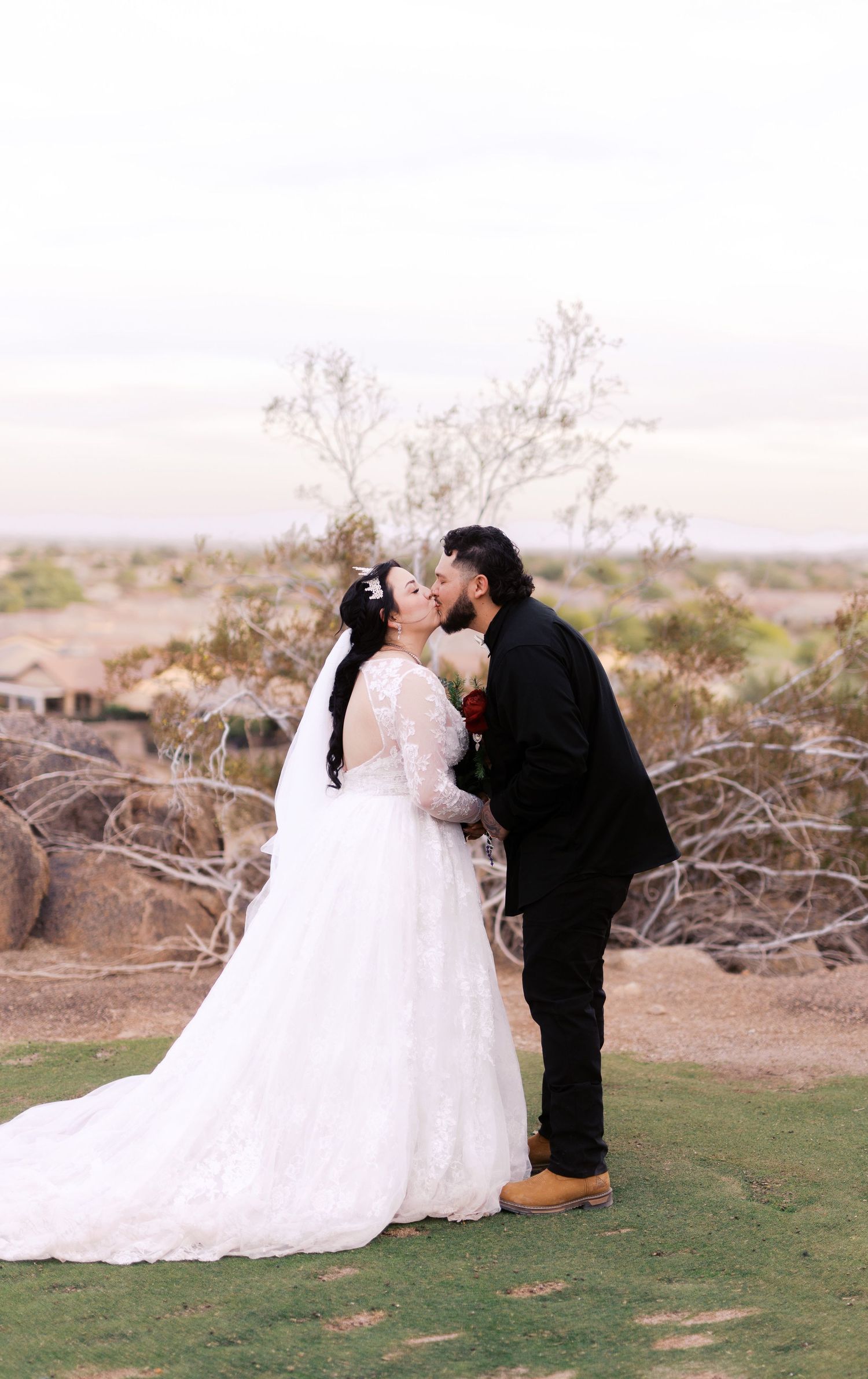 A couple in wedding attire share a romantic moment on a grassy overlook with desert landscape in the background.