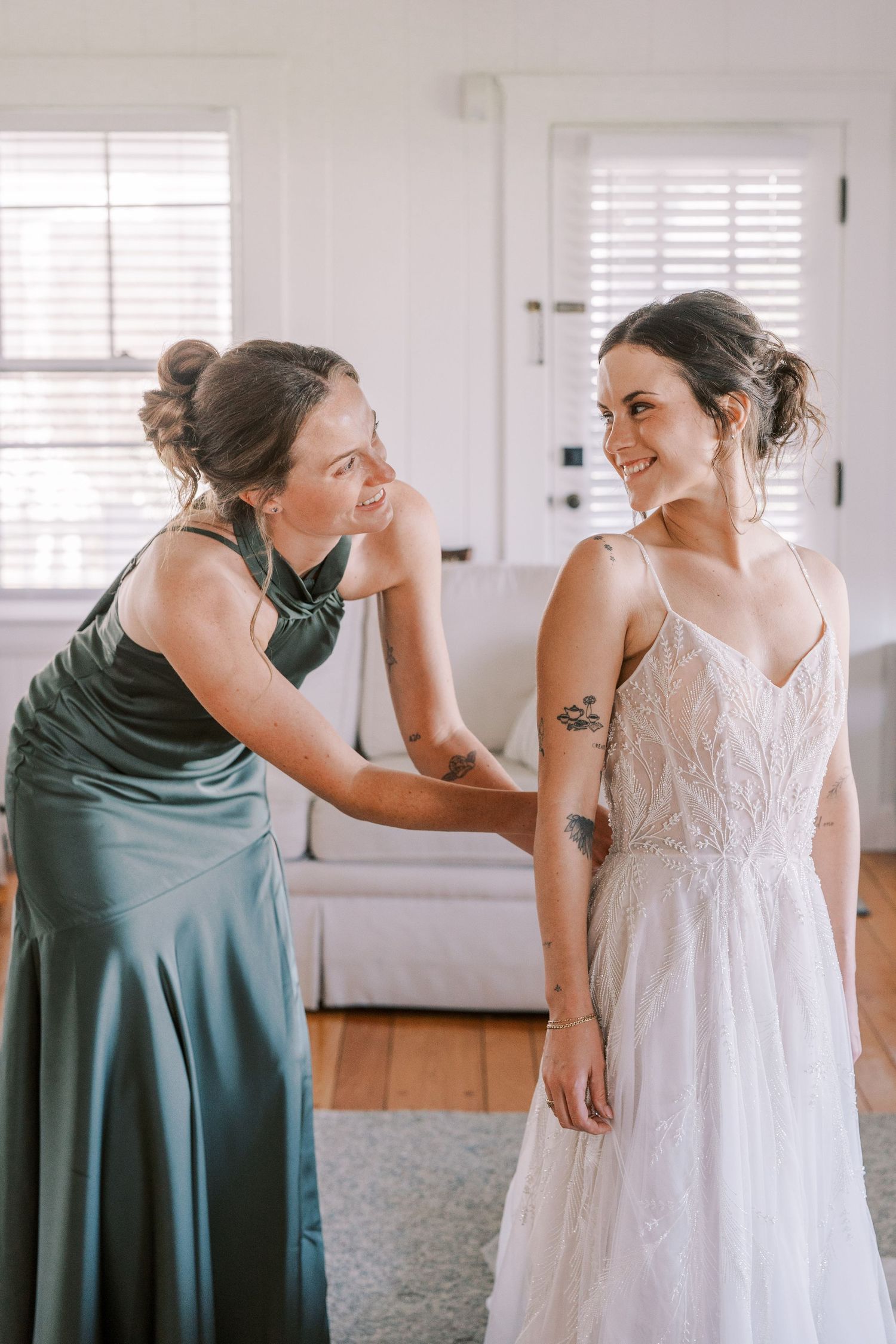 Getting ready moment with bridesmaid helping adjust wedding dress in bright room.
