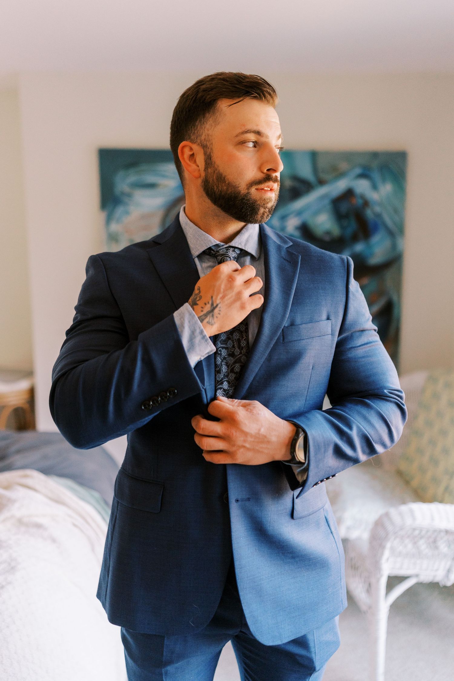 Man in navy blue suit jacket adjusts his collar and tie while getting dressed in a bright interior setting.