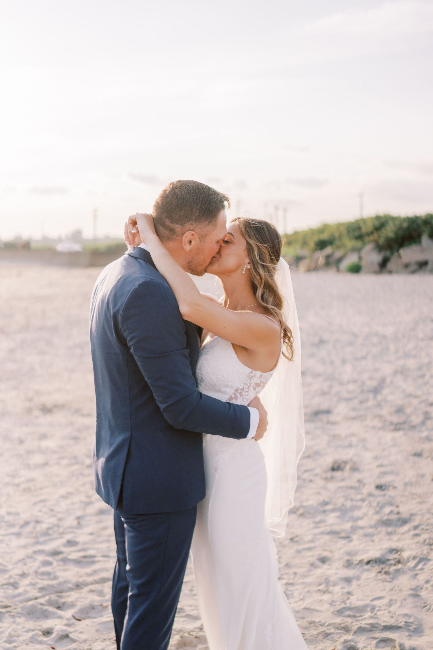 A romantic couple shares a tender kiss on a sandy beach at sunset while wearing elegant wedding attire.