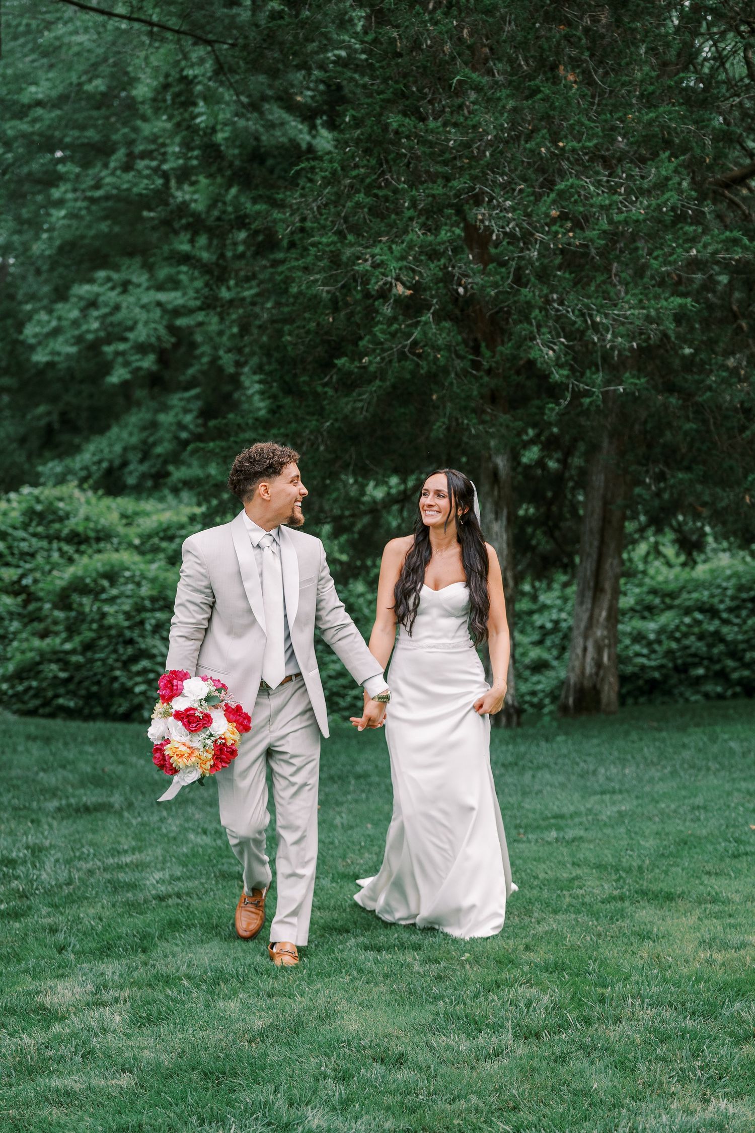 Newlyweds walk hand in hand across a lush green lawn with the bride holding a vibrant red and white bouquet.