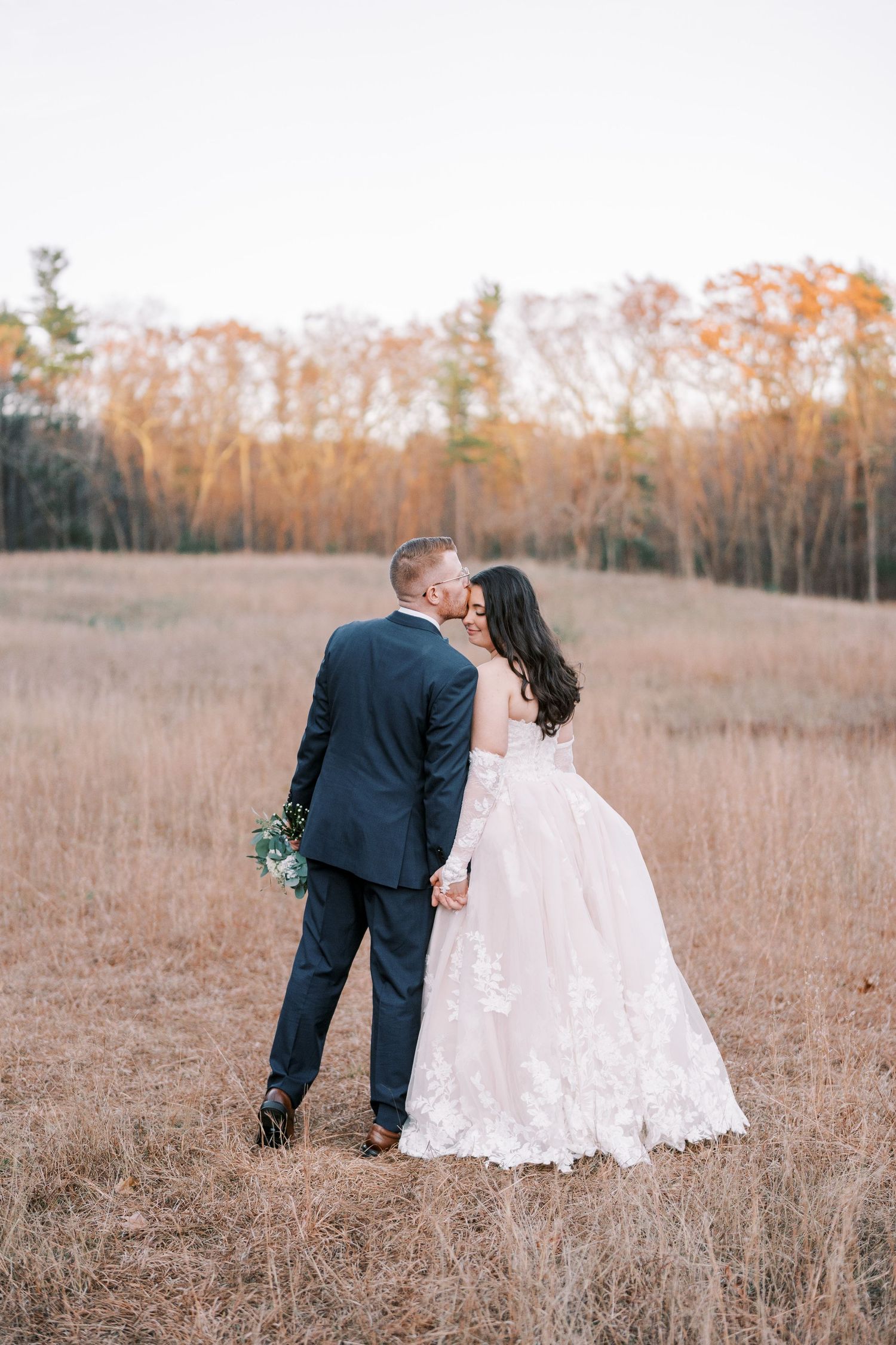A couple in wedding attire share an intimate moment in a golden autumn field during their outdoor photoshoot.