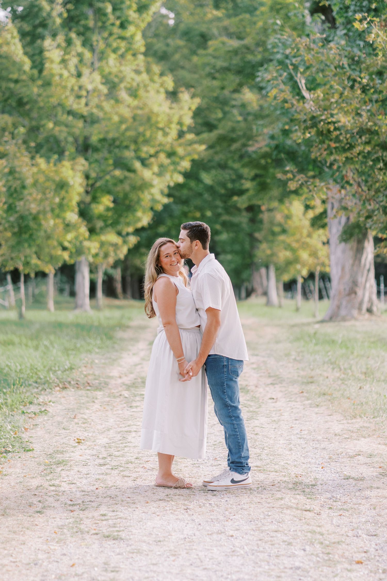 Romantic couple embracing on a tree-lined path in casual summer attire.