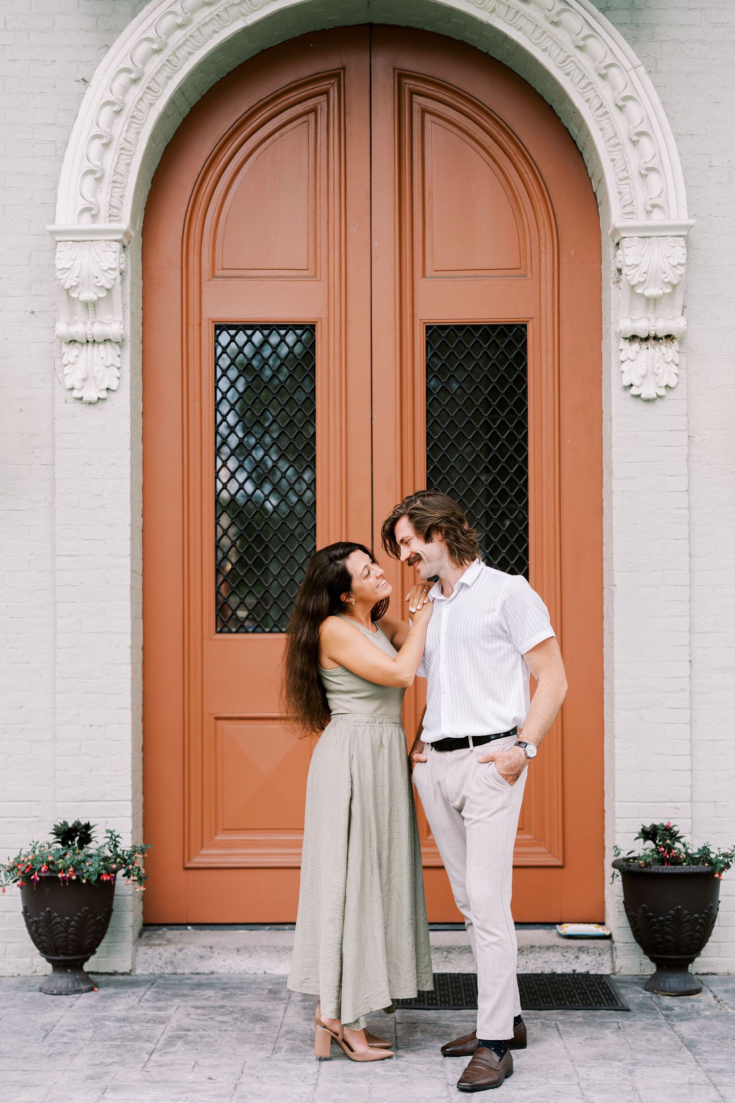 A couple shares a romantic moment in front of large coral-colored doors with ornate white architectural details.