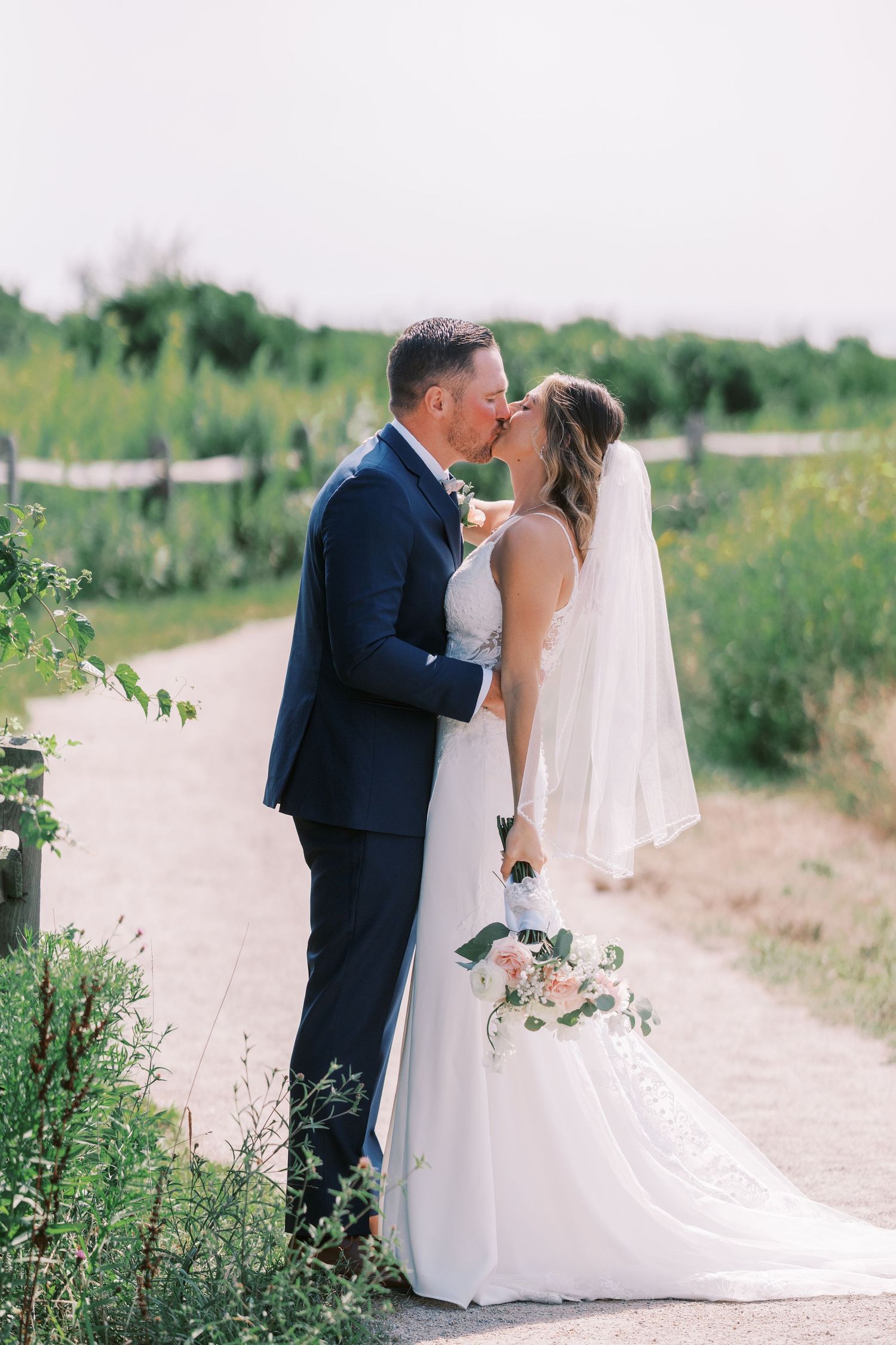 A romantic wedding couple shares intimate moments on a rustic dirt path surrounded by summer greenery.