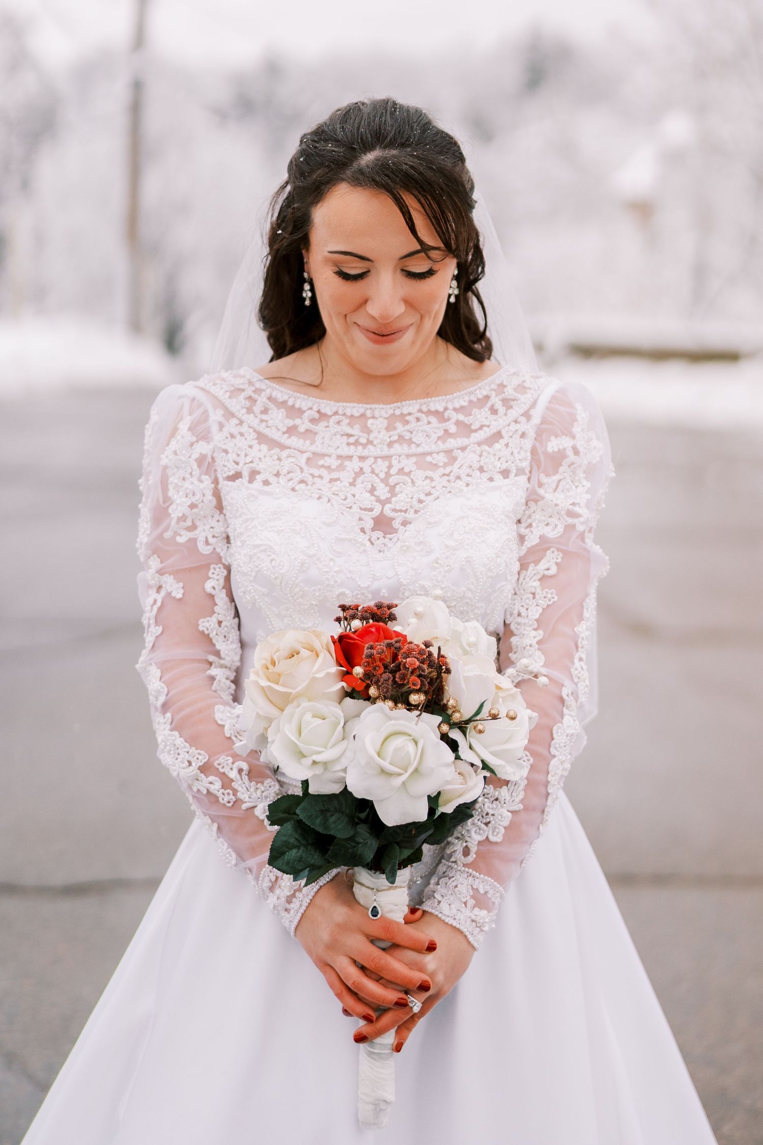 Bride in long sleeve lace wedding dress holding white and red bouquet against snowy backdrop.