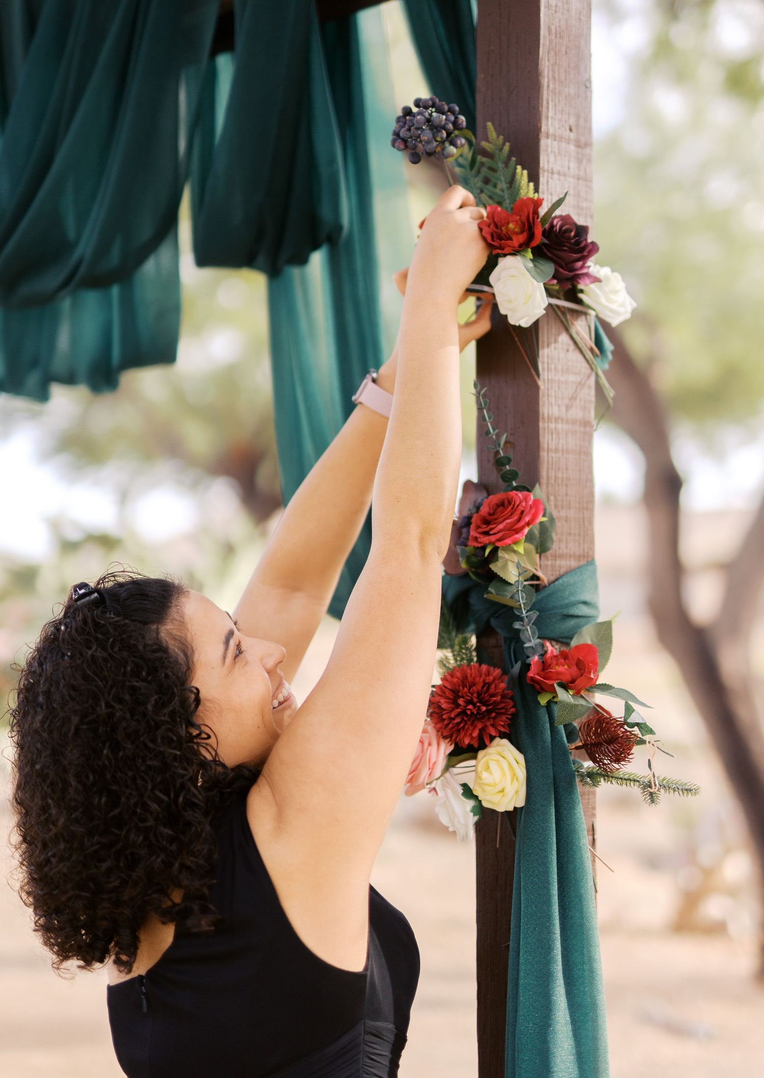 Event decorator arranging beautiful red and white flowers on a teal draped wedding arch outdoors.