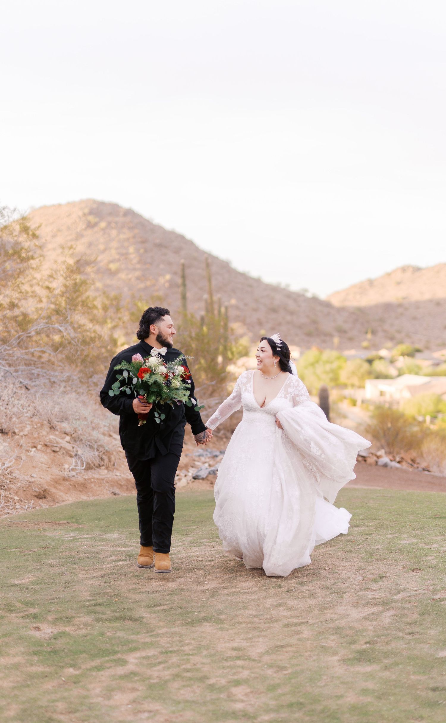 A couple in wedding attire walks together on a grassy hill with desert mountains and cacti in the background.