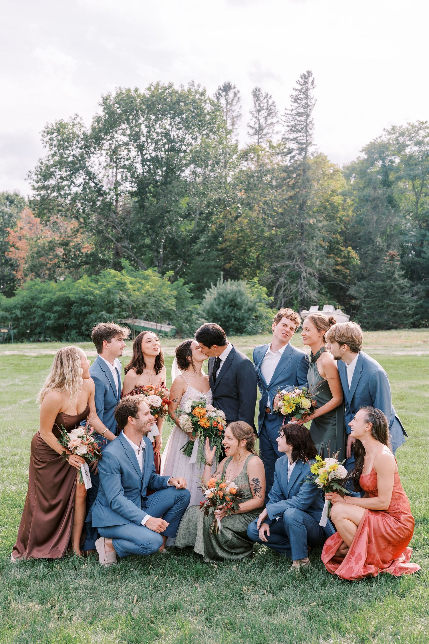 Wedding party lounges on grass in coordinated attire of navy suits and earth-toned dresses.