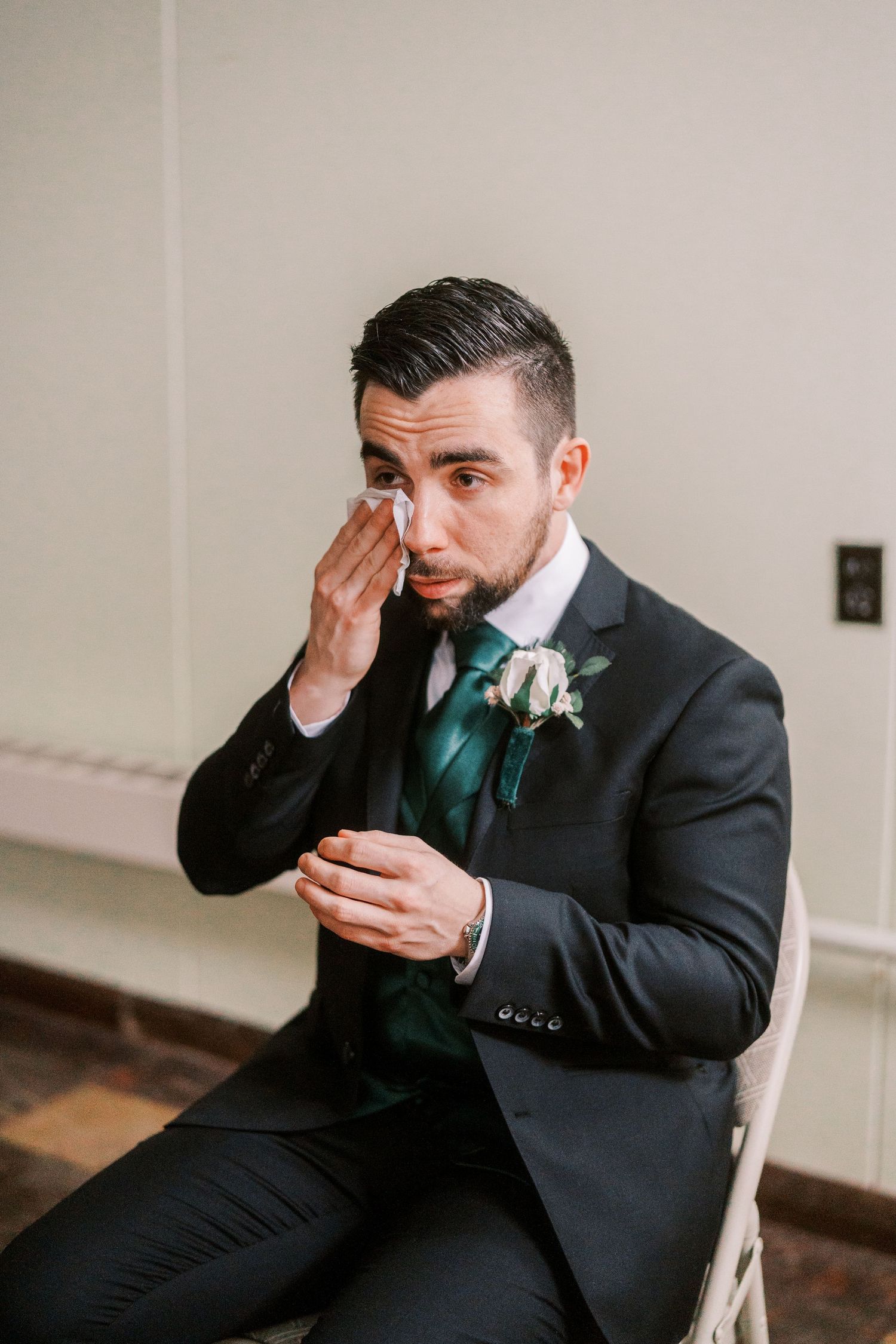 An emotional groom in a black suit and green tie wipes tears from his eyes.