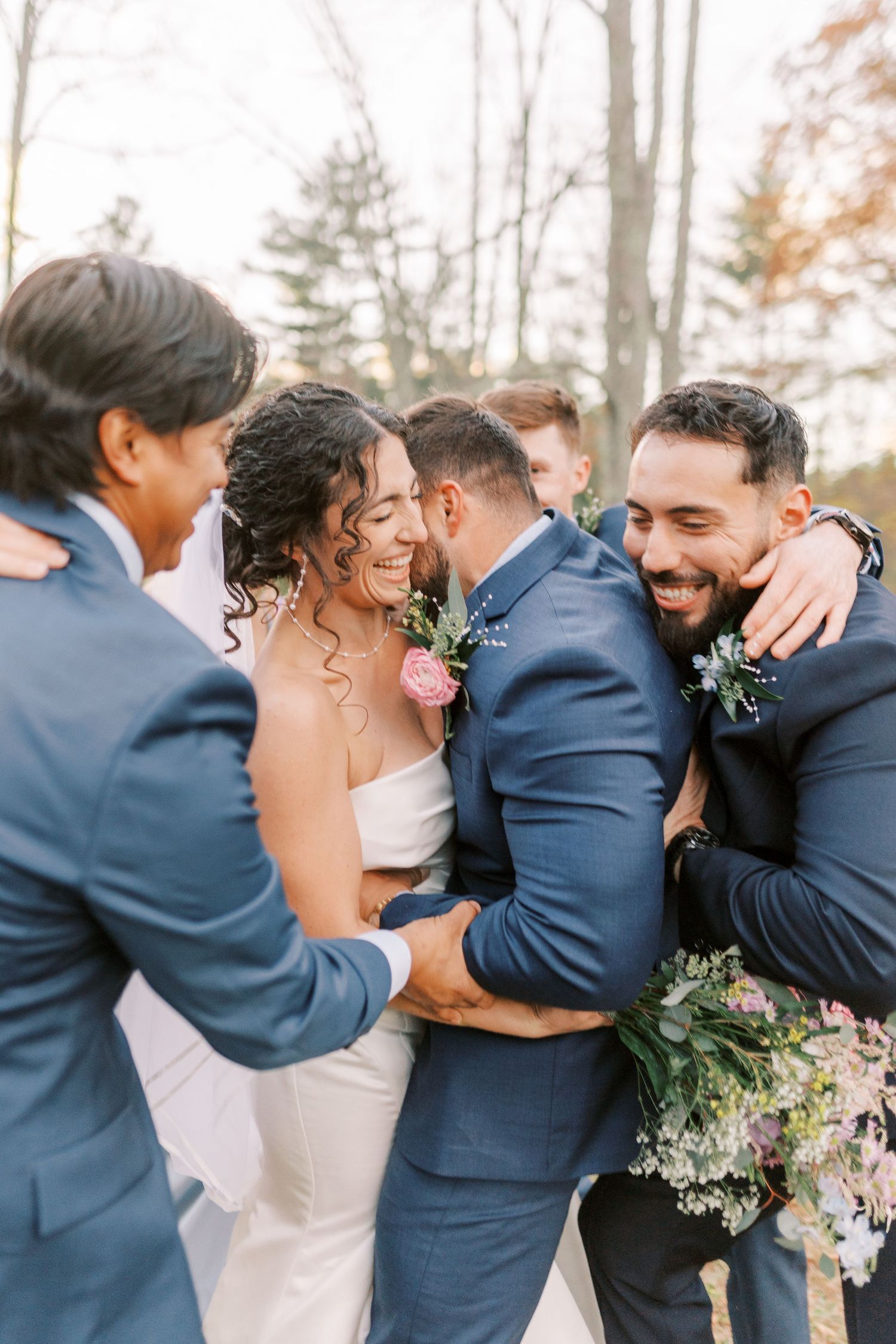 Wedding party shares a joyful embrace outdoors wearing navy blue suits and holding floral bouquets.