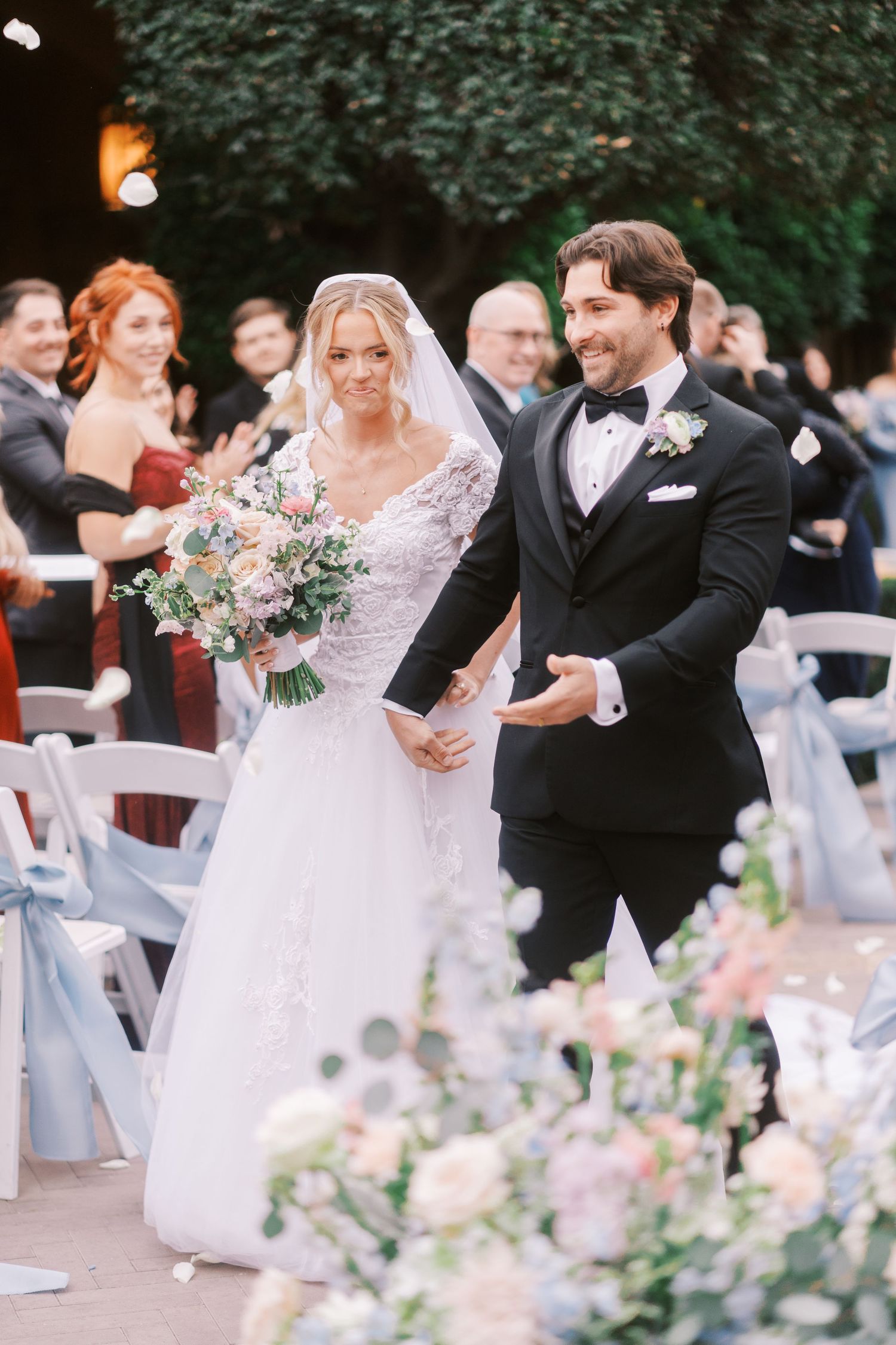 Wedding party dances and celebrates during outdoor garden reception with pink floral centerpieces and white chairs.