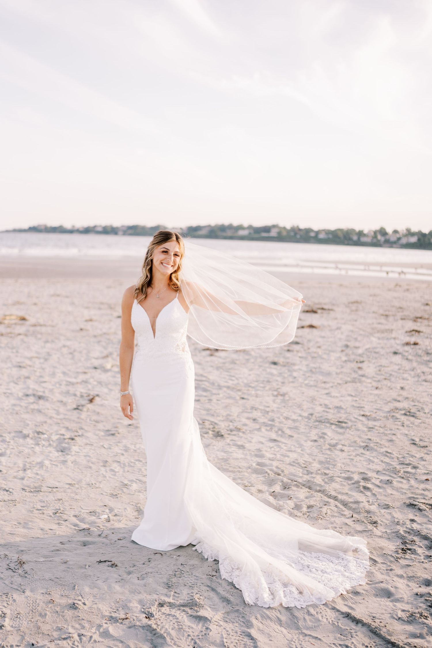A bride in a flowing white wedding dress poses gracefully on a sandy beach during sunset.