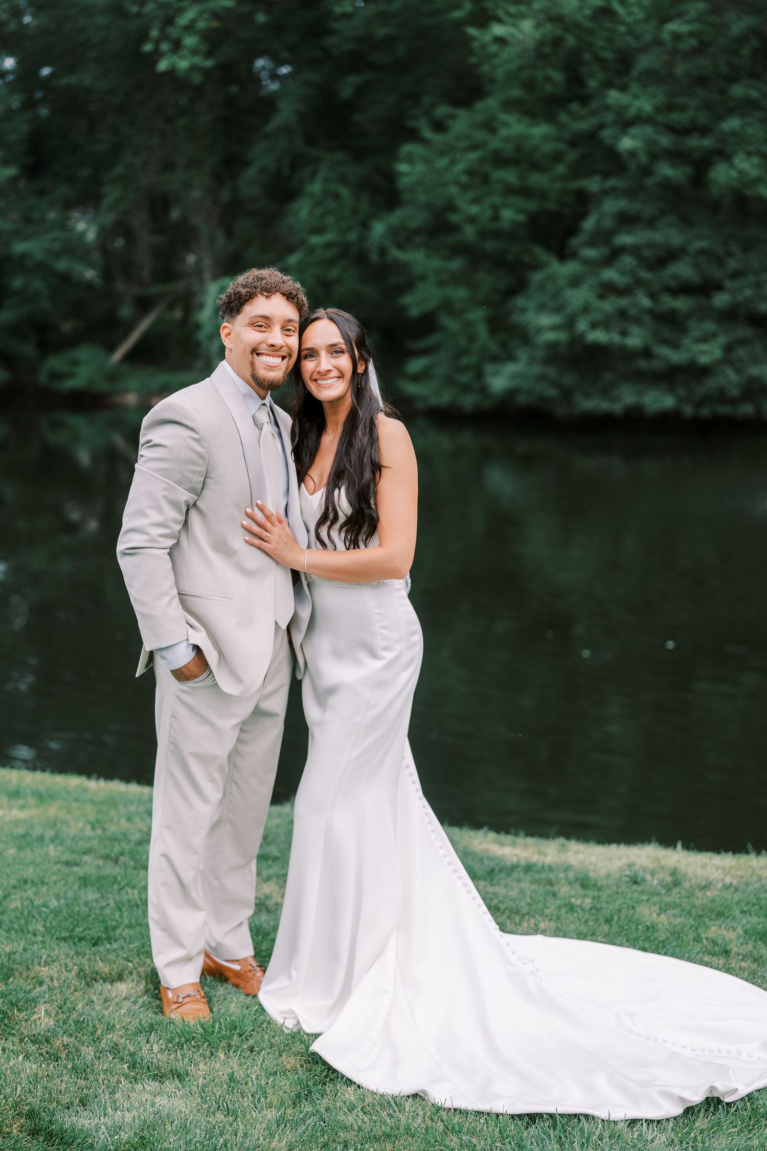 Newlyweds pose together on lawn with lake and trees in background.