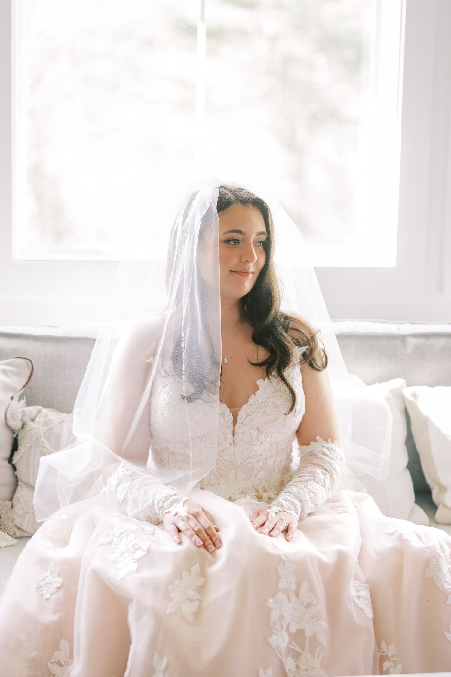 Bride in white lace wedding dress sits gracefully on light-colored couch in bright sunlit room.