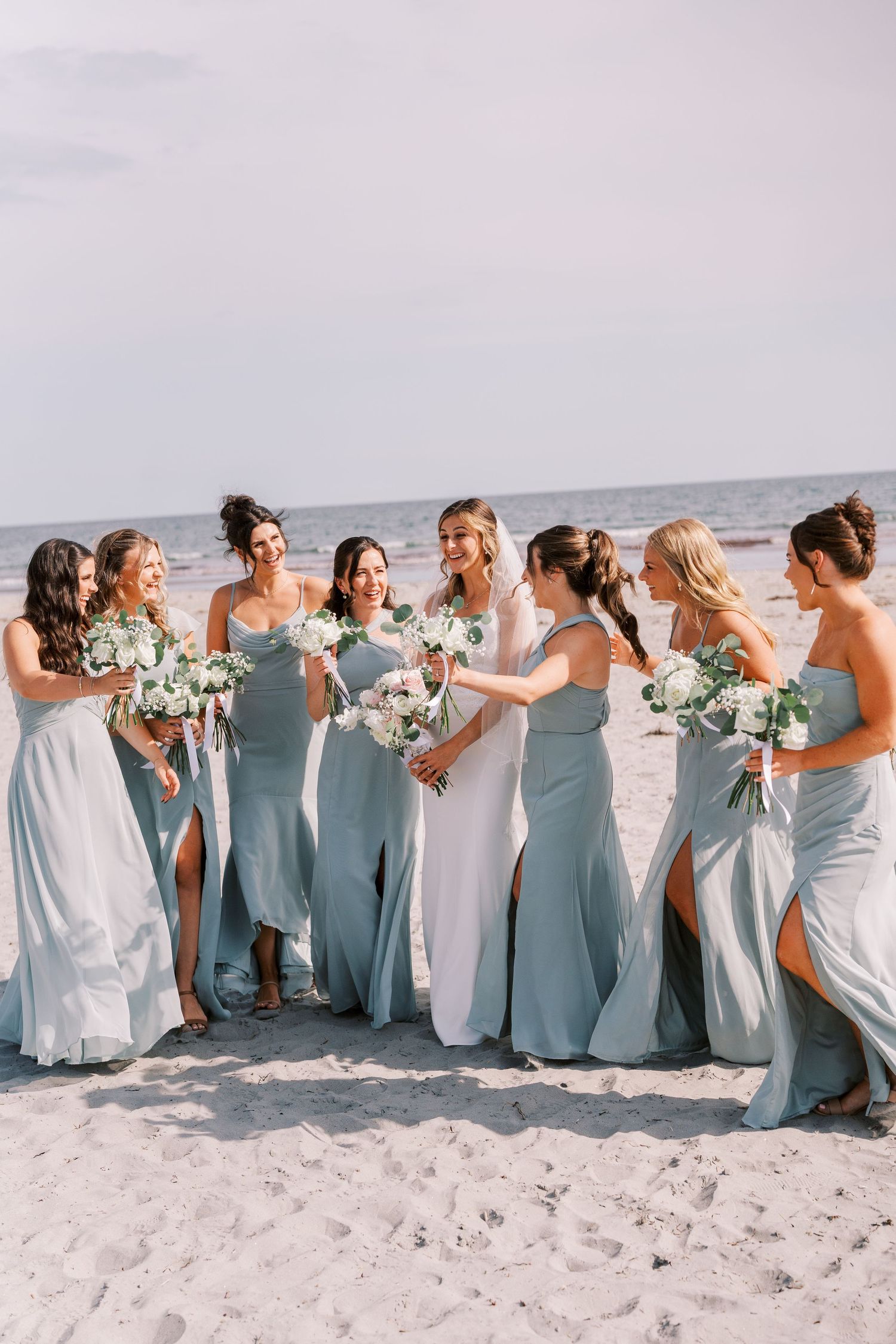 A bridal party in light blue and white dresses laughs together while standing in a line on a sunny beach.