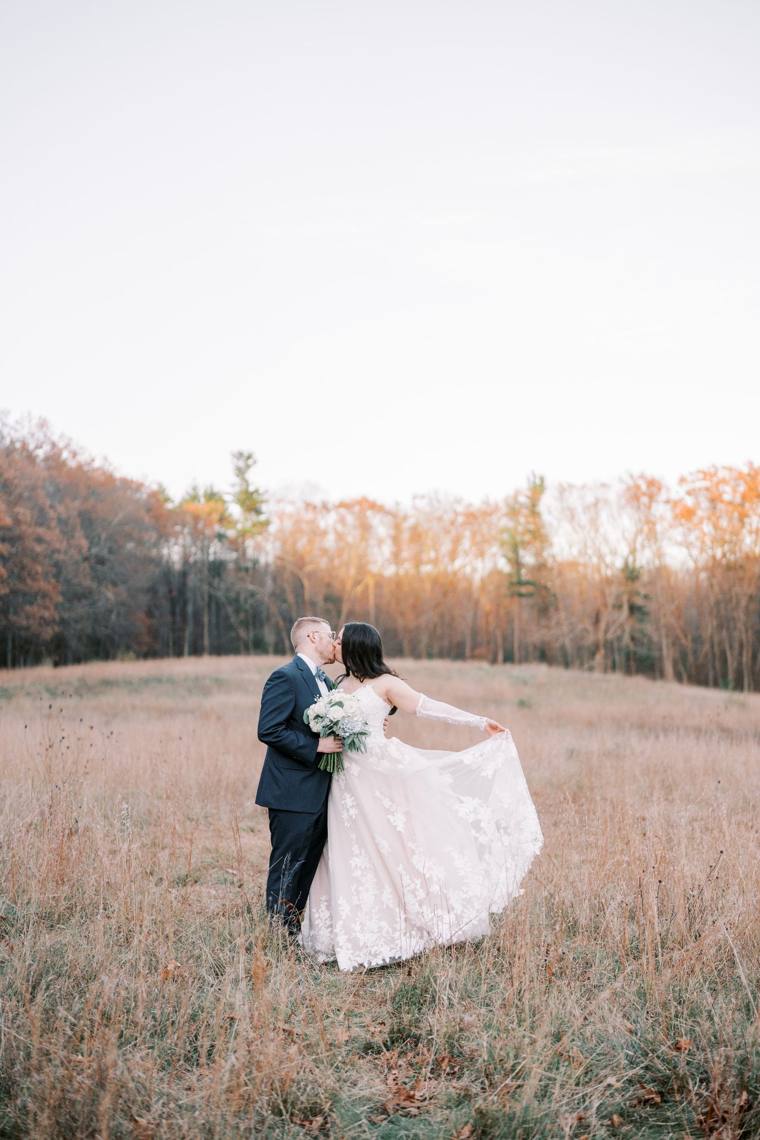 A romantic wedding couple embraces in a golden autumn field at sunset as the bride's lace dress flows in the breeze.