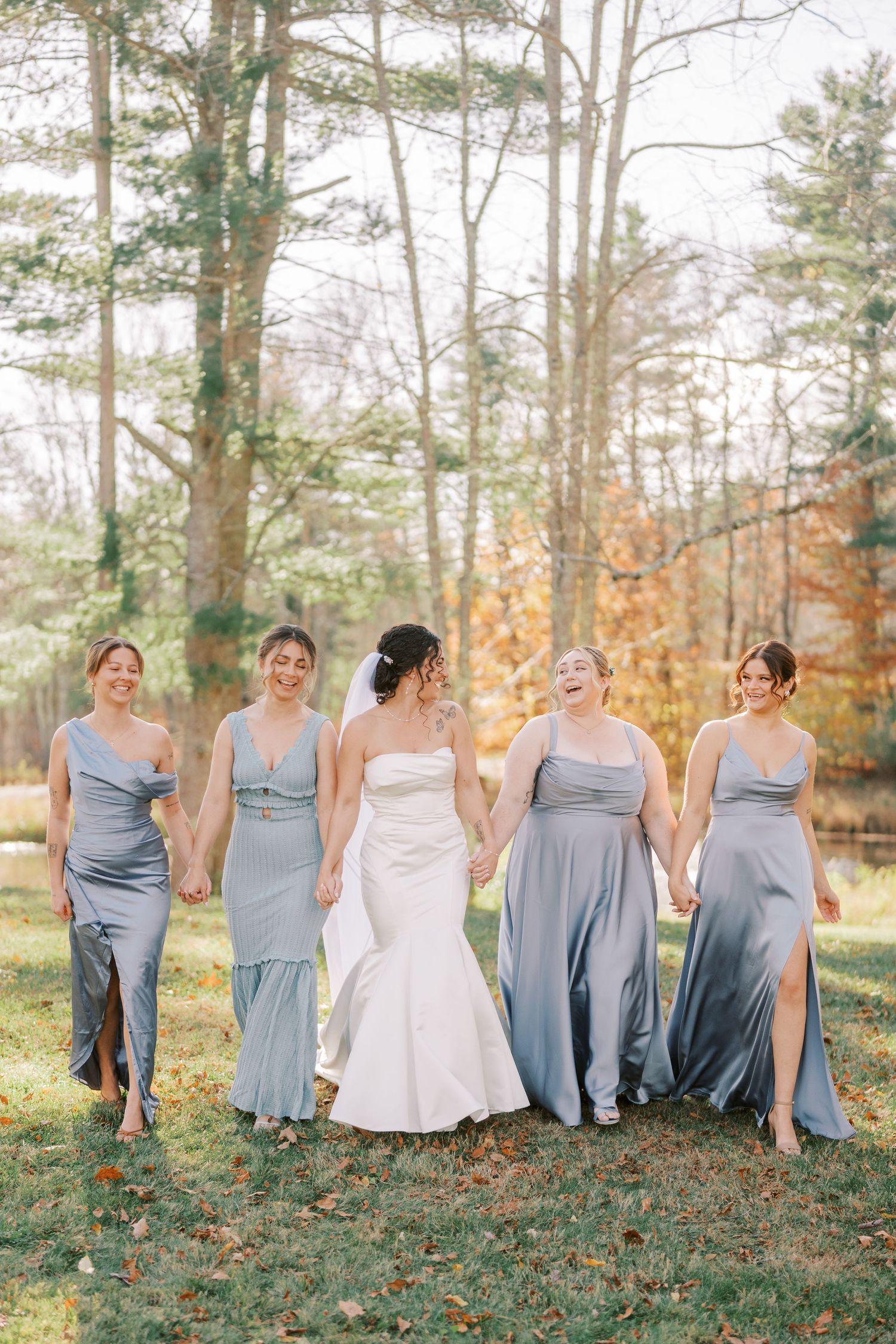 A bridal party in dusty blue dresses and white gown poses together in a scenic autumn forest setting.