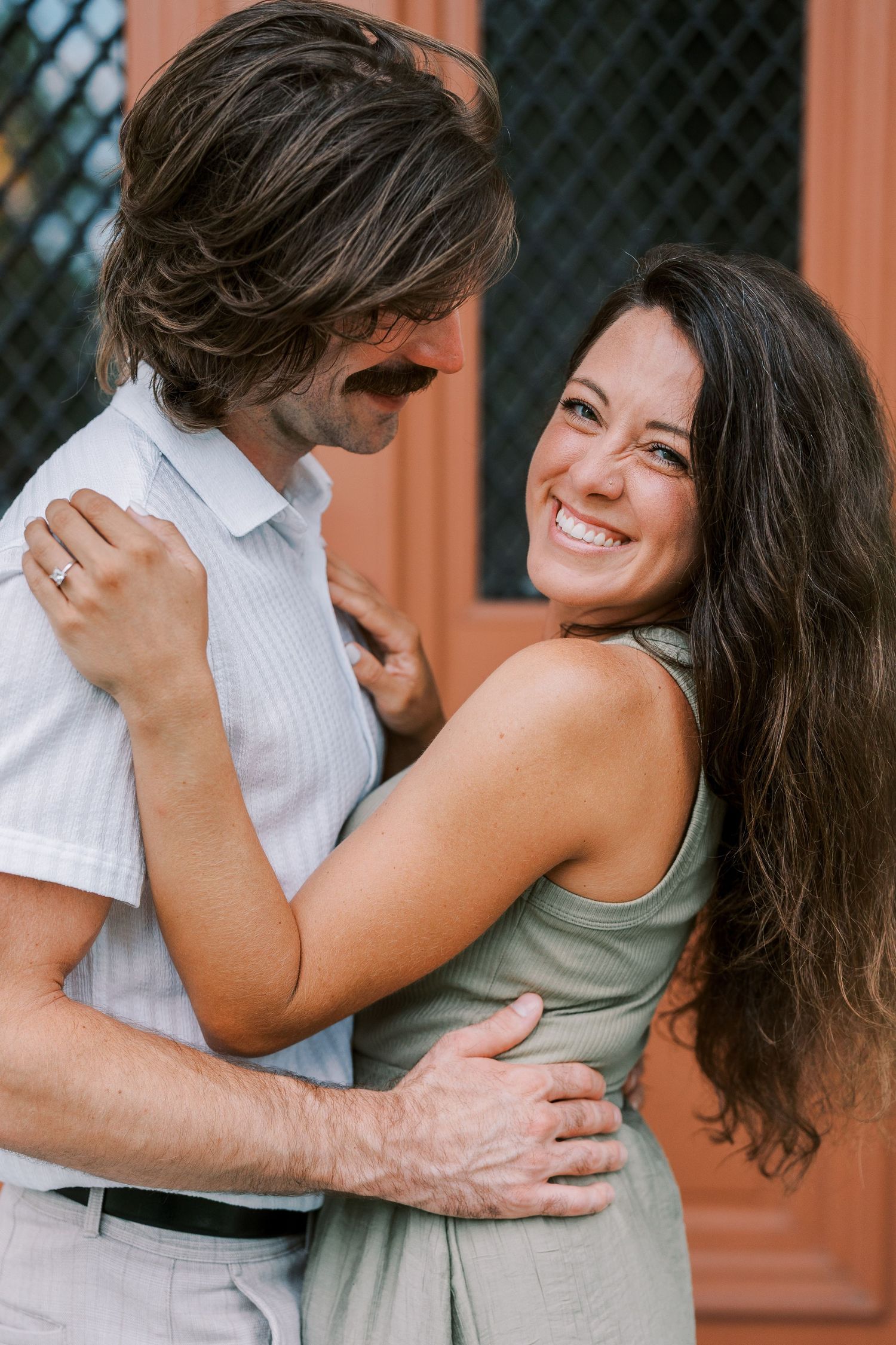 A romantic couple in white and olive outfits embracing and laughing against a terracotta wall.