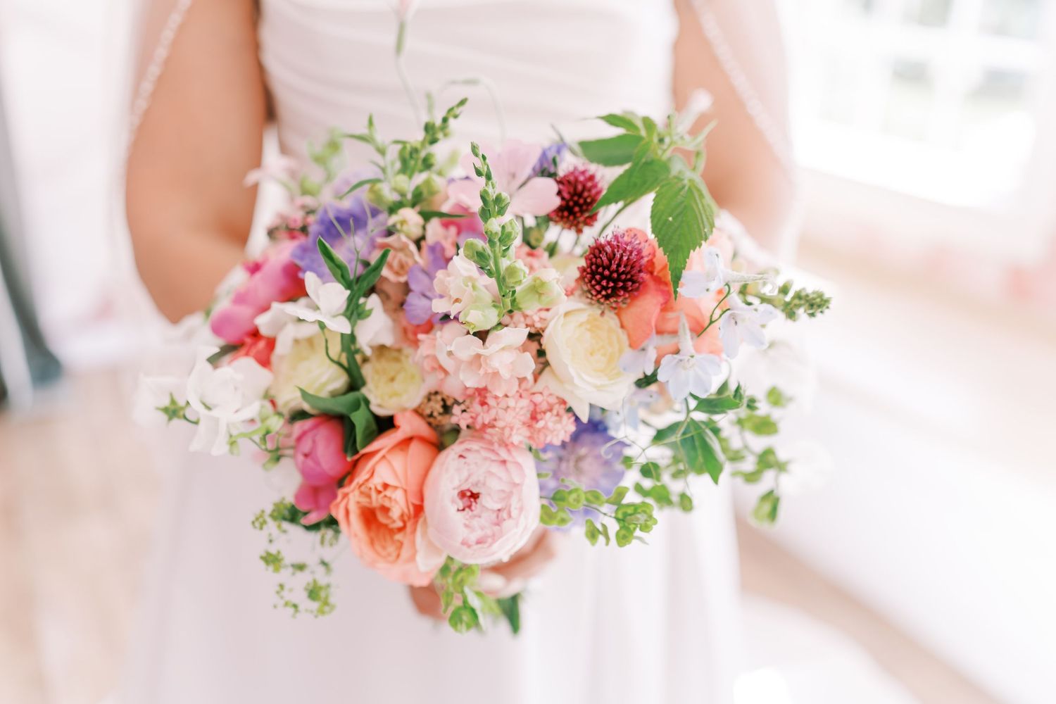 A romantic wedding bouquet featuring pink peonies, coral roses, purple blooms and delicate greenery in soft lighting.