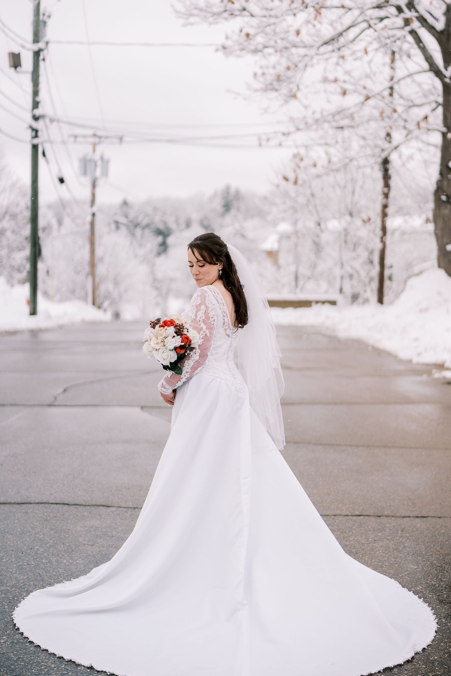 Bride in lace-sleeved wedding gown with long train poses on snowy winter street.