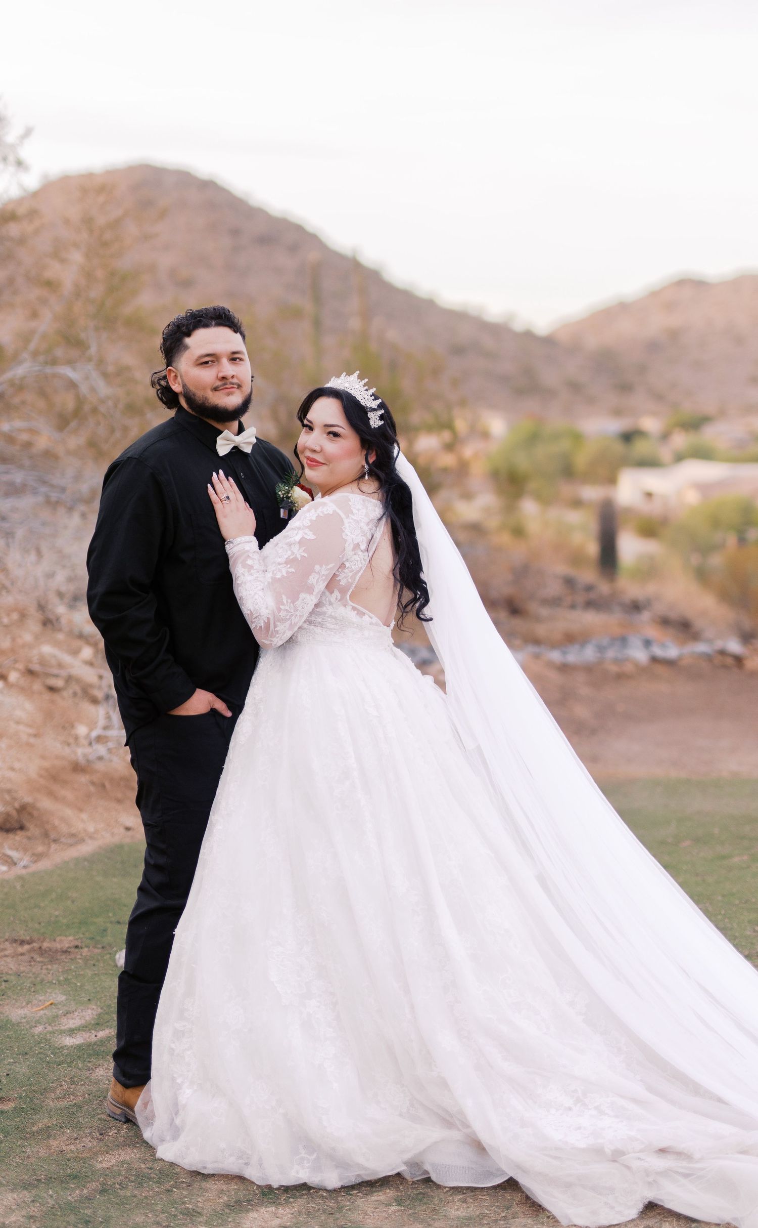 A bride in a flowing white dress and veil stands with her groom against a desert mountain backdrop during golden hour.