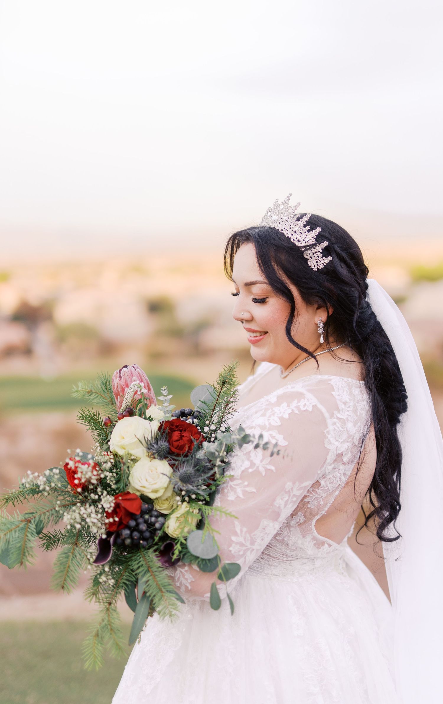 A bride in a white lace dress and crystal crown holds a bouquet featuring red and white flowers with greenery.