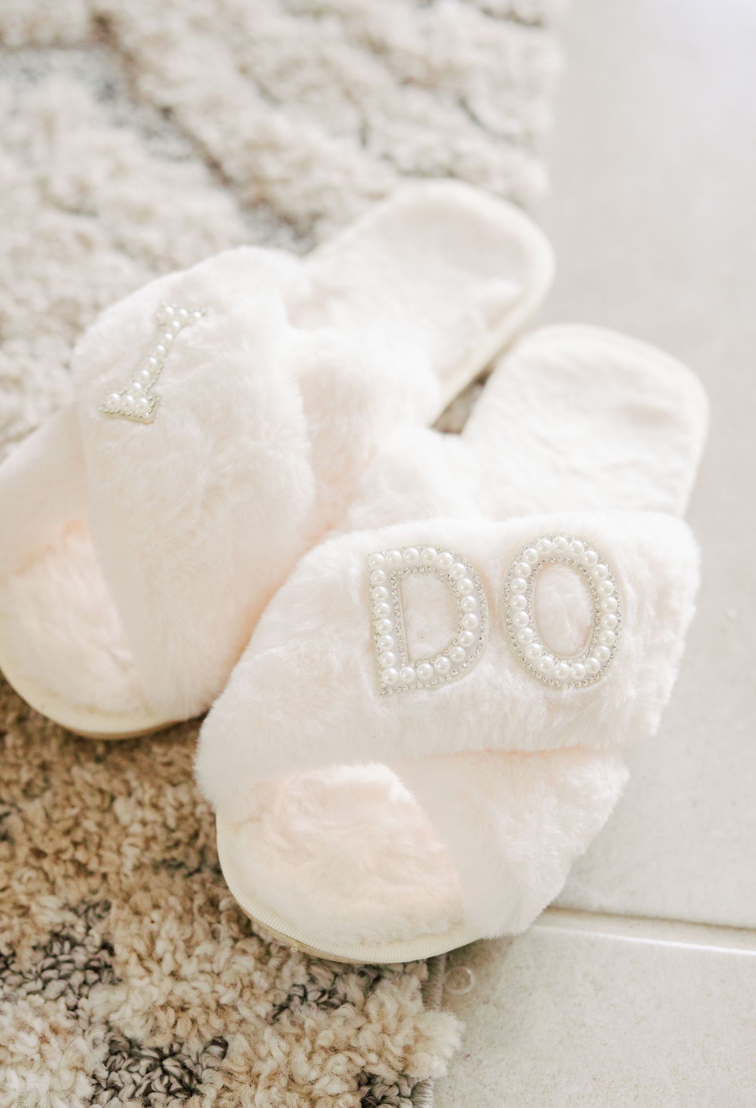 White plush slippers with beaded 'I DO' text on top sitting on marble floor, perfect for a bride's wedding day.