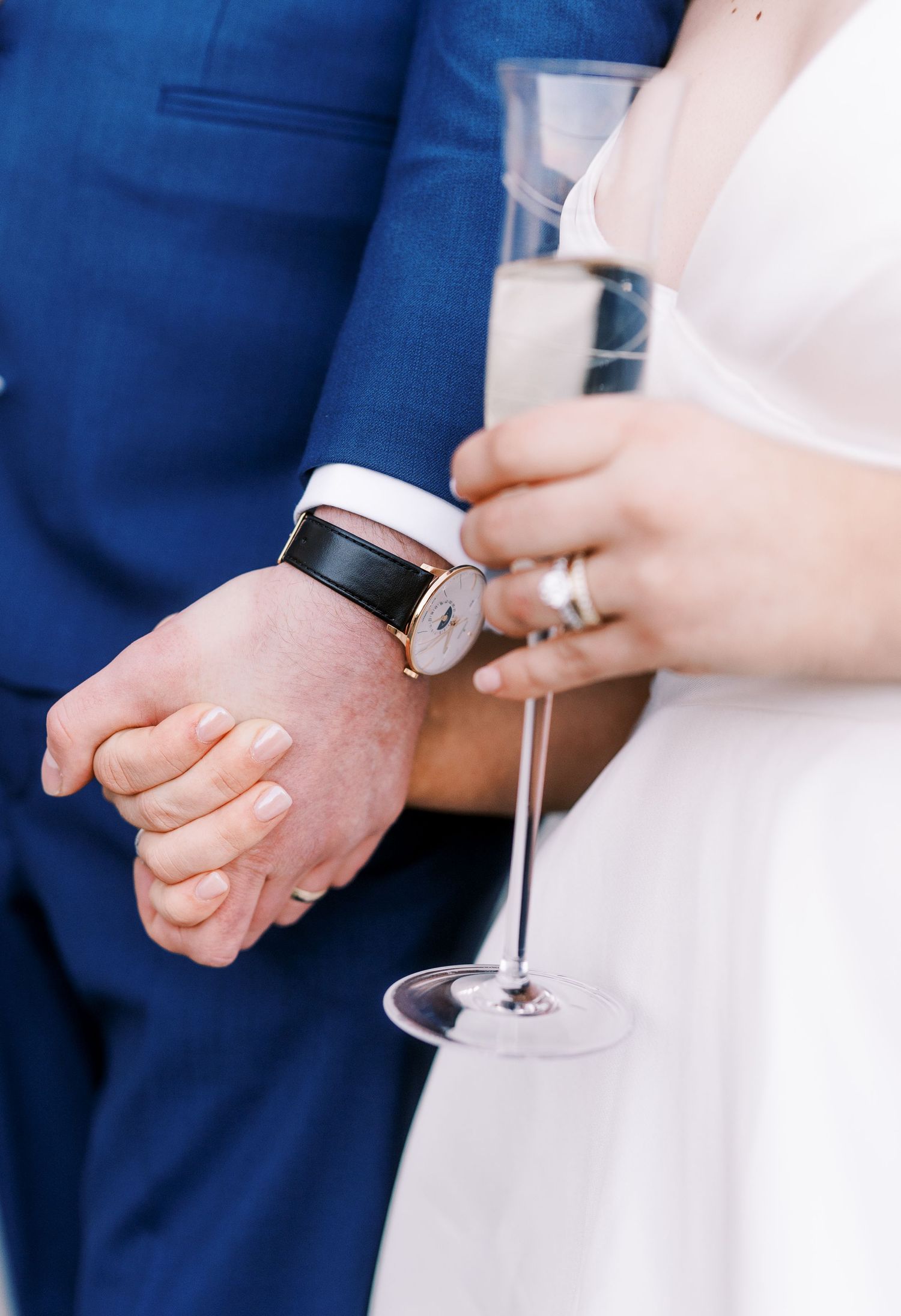 Close-up of champagne glass and smart watch detail during wedding celebration with blue suit and white dress.