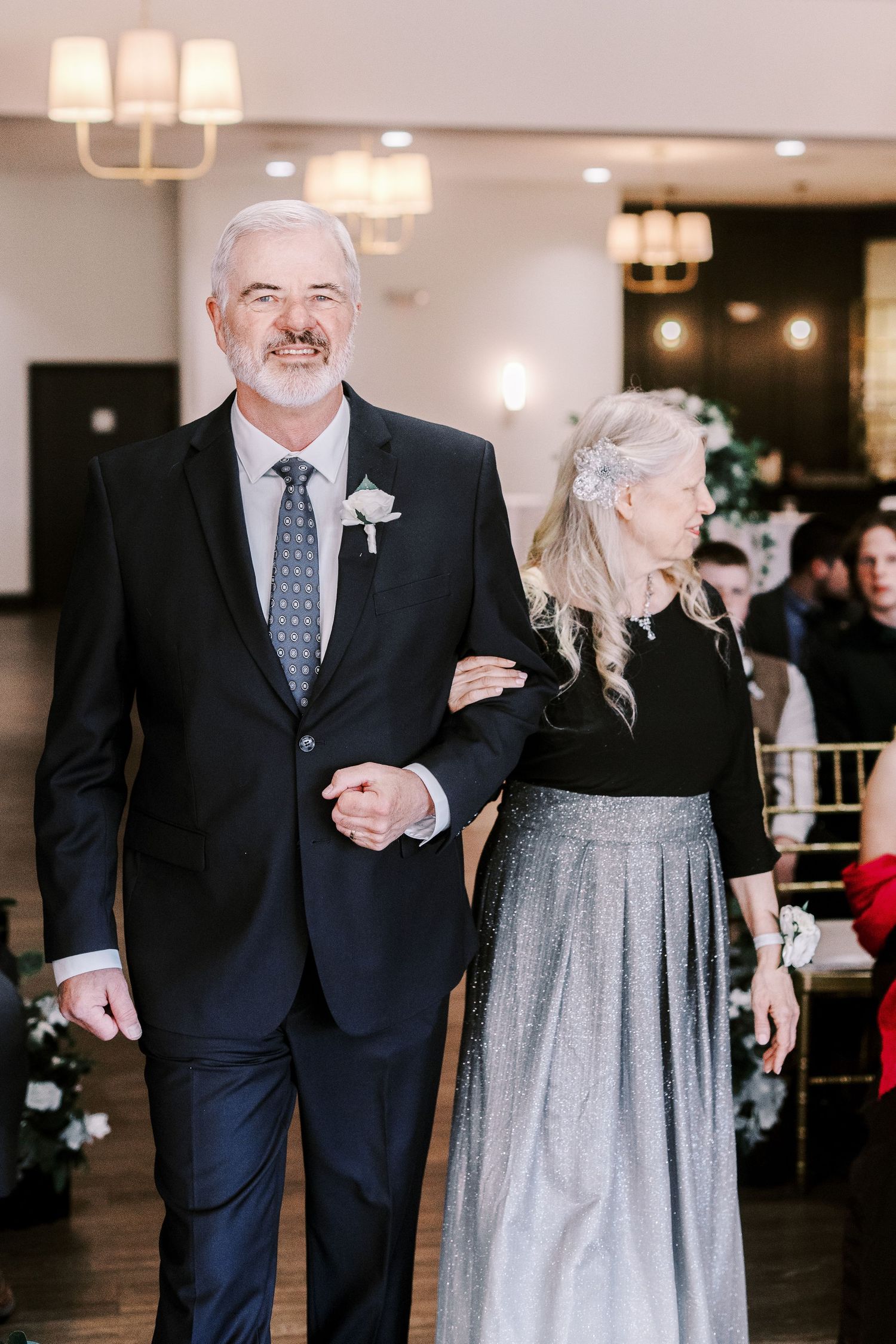 A couple in formal attire walk arm-in-arm at a wedding event, with the woman wearing a sparkling silver skirt.