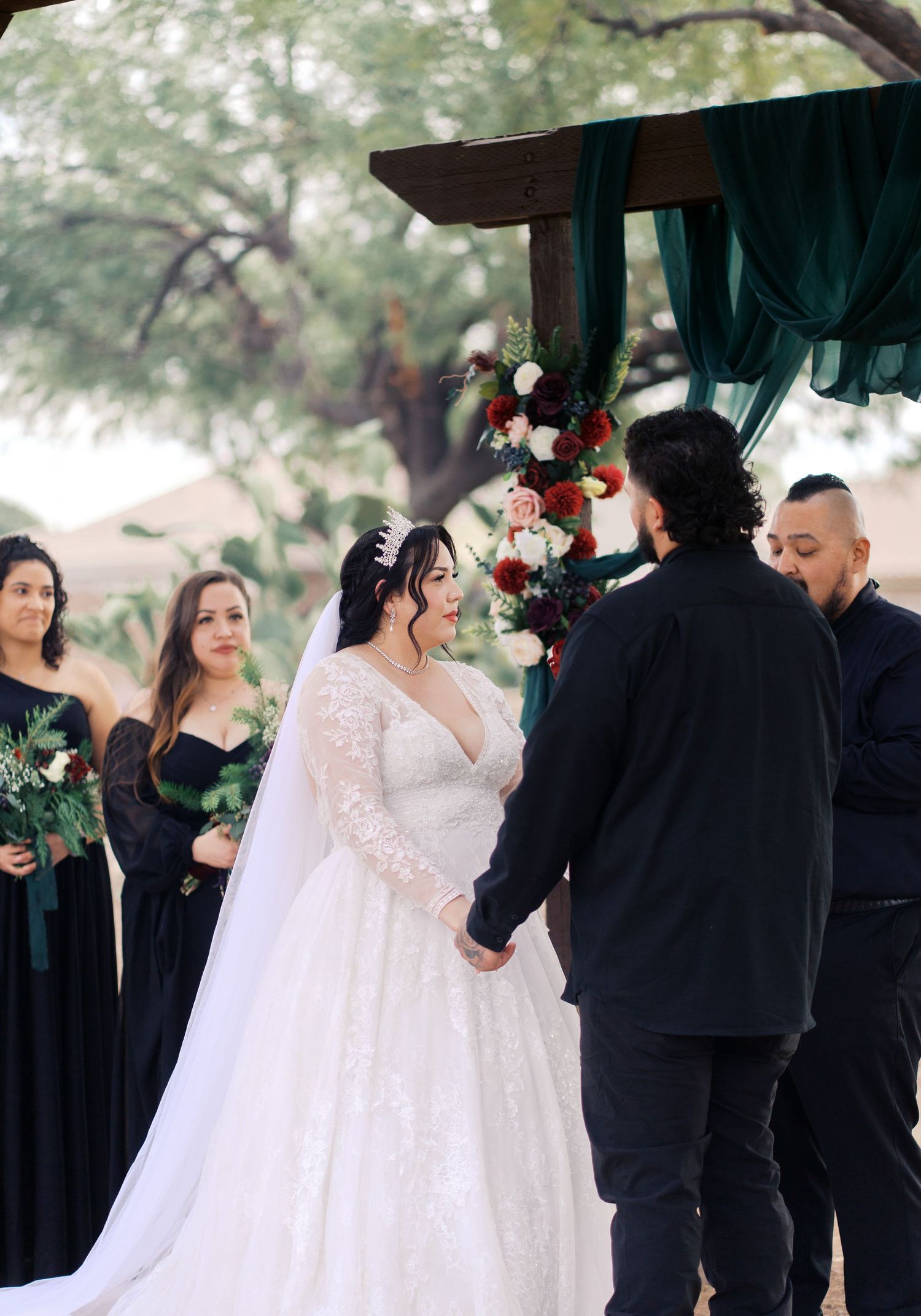 A romantic outdoor wedding ceremony under a wooden arch decorated with flowers and green fabric draping.